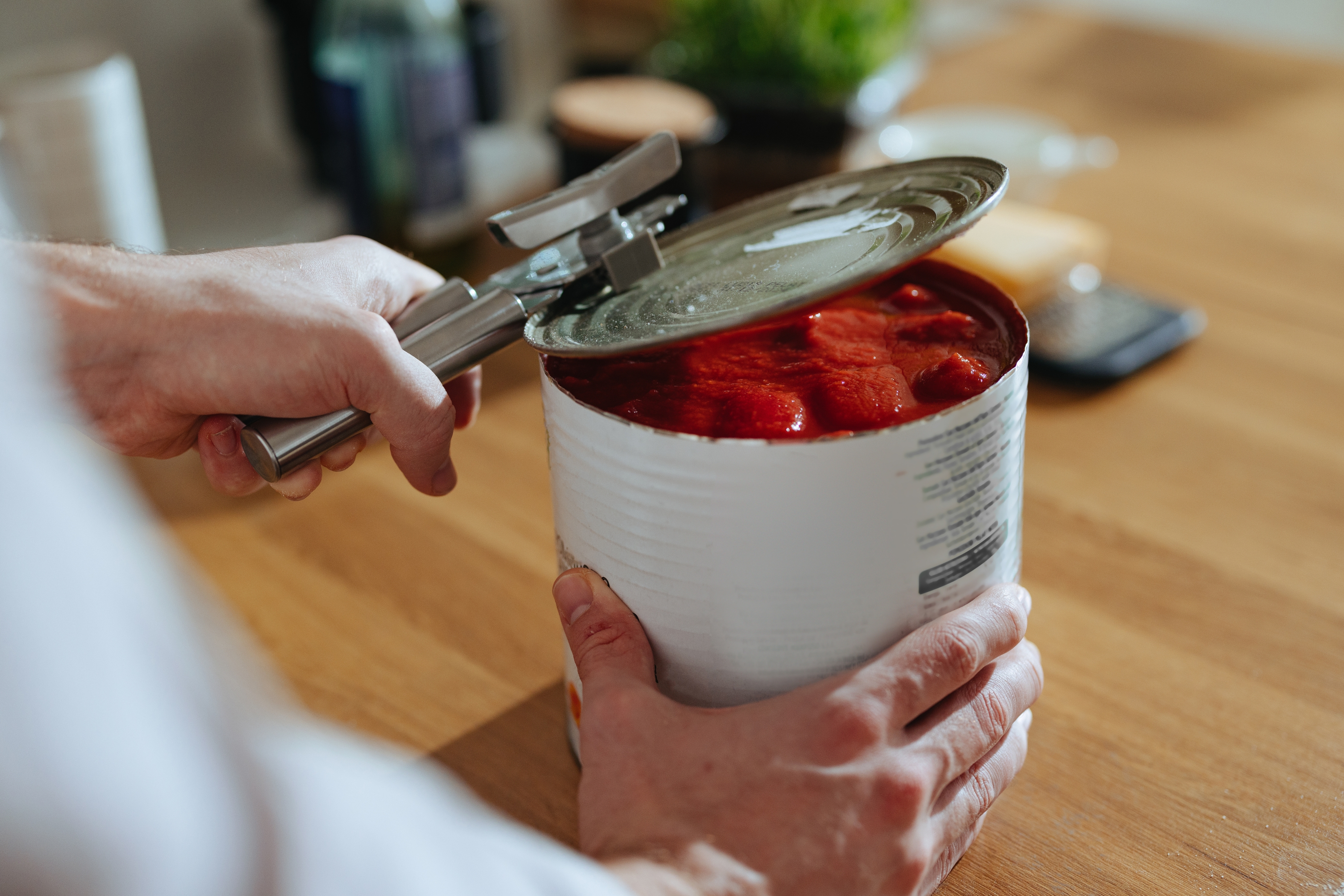 Person opening a can of whole peeled tomatoes with a can opener on a wooden table