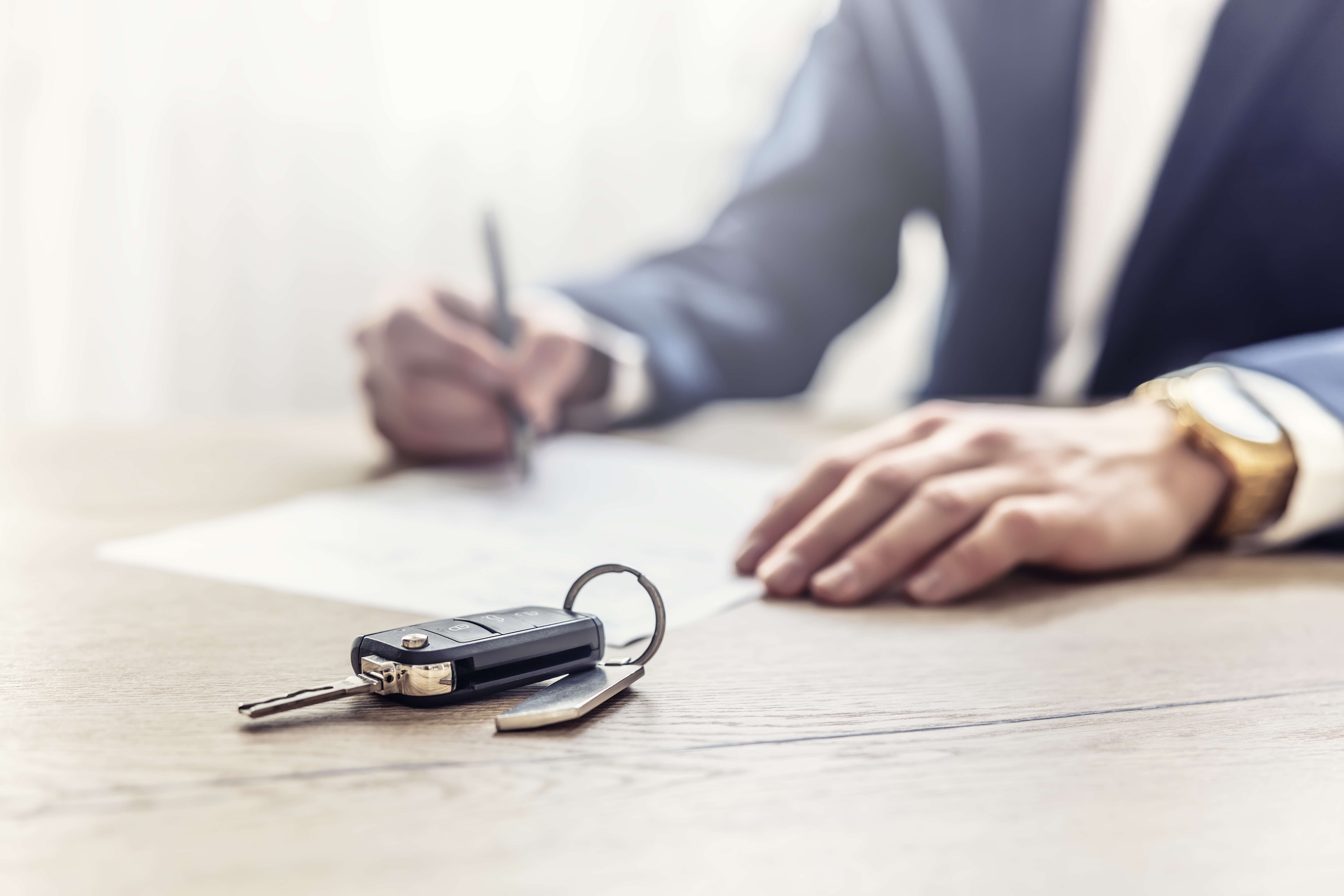 A person in a suit signs papers on a table, with car keys in the foreground