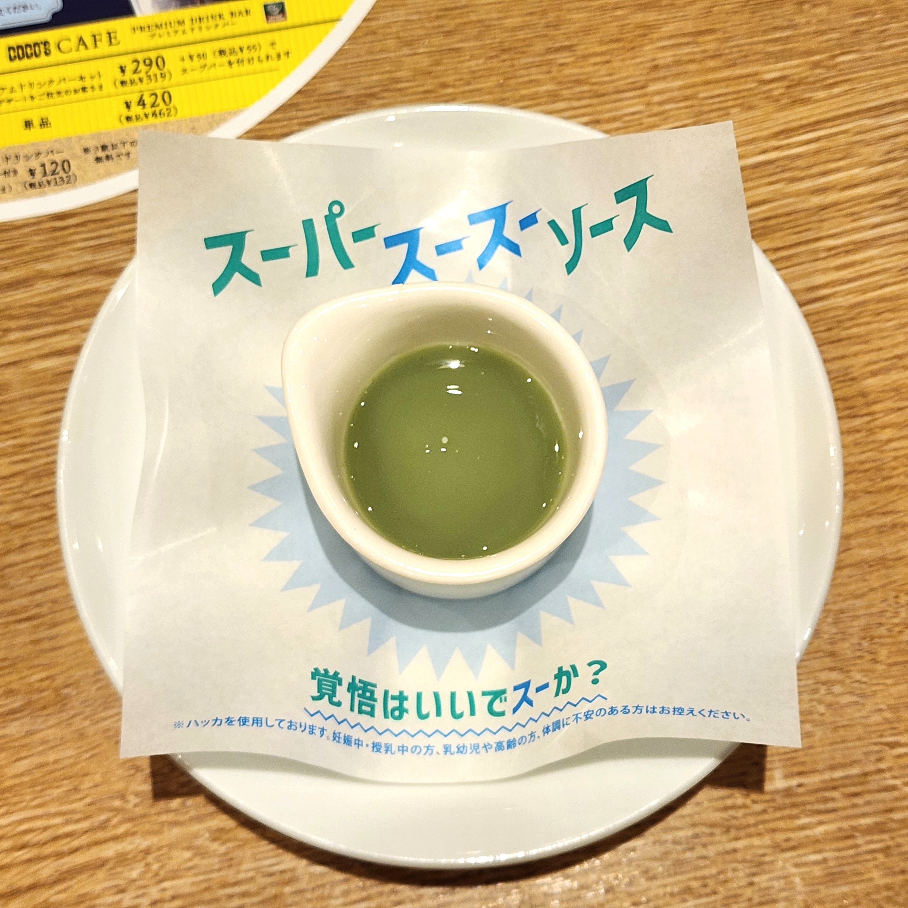 緑色のソースが入った白い小皿。紙には「スーパーソースソース」と書かれており、キャッチフレーズが下部に記載されています。