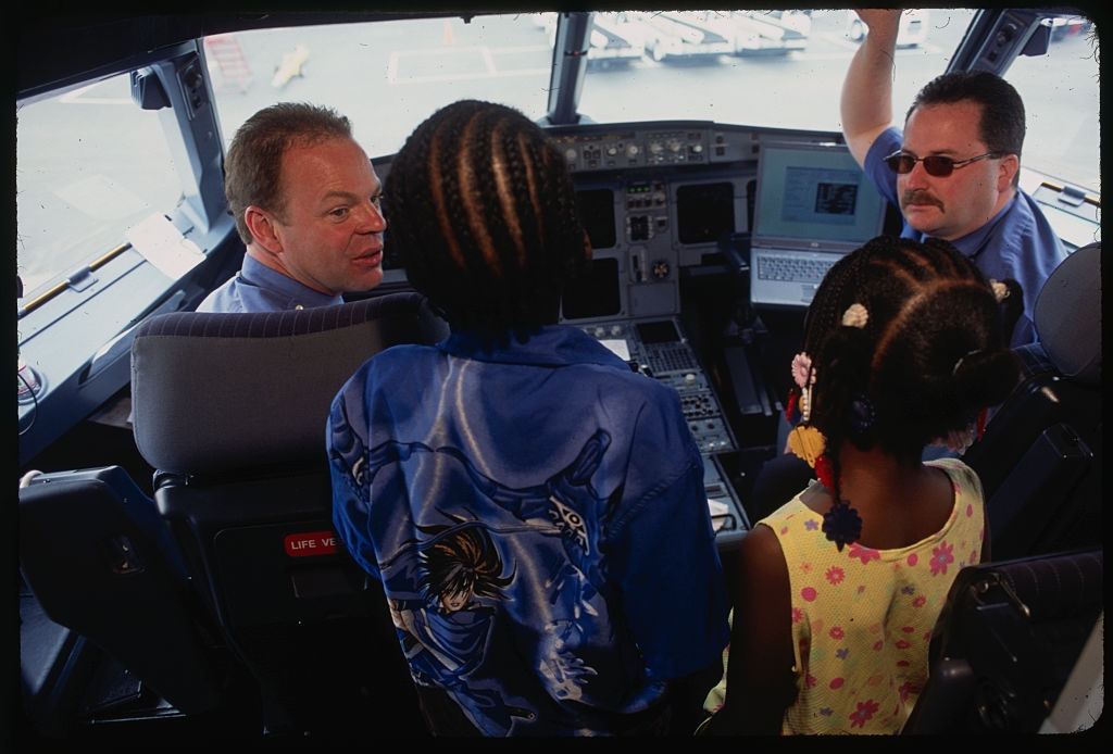 Two pilots interact with two kids in a cockpit; one child wears a patterned shirt, and the other has beaded hair, observing the airplane controls