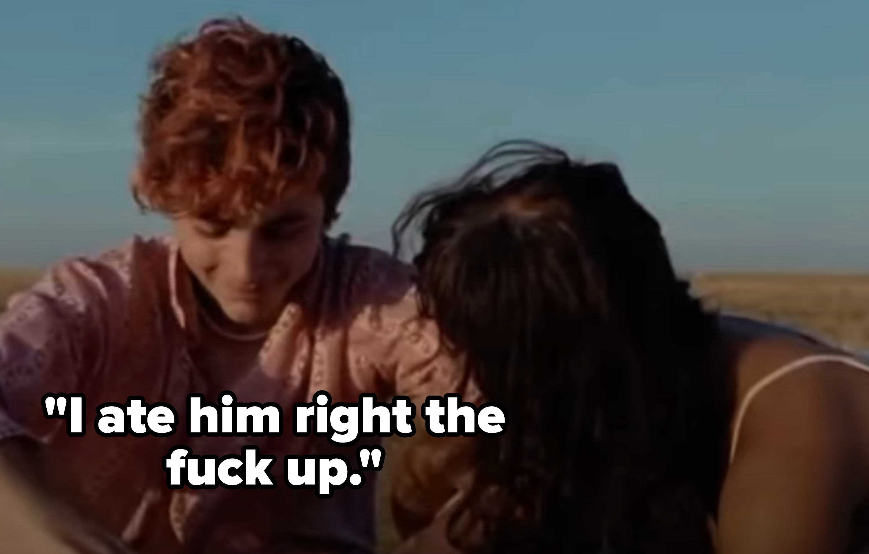 Two people seated outdoors, smiling and leaning close, with a field in the background