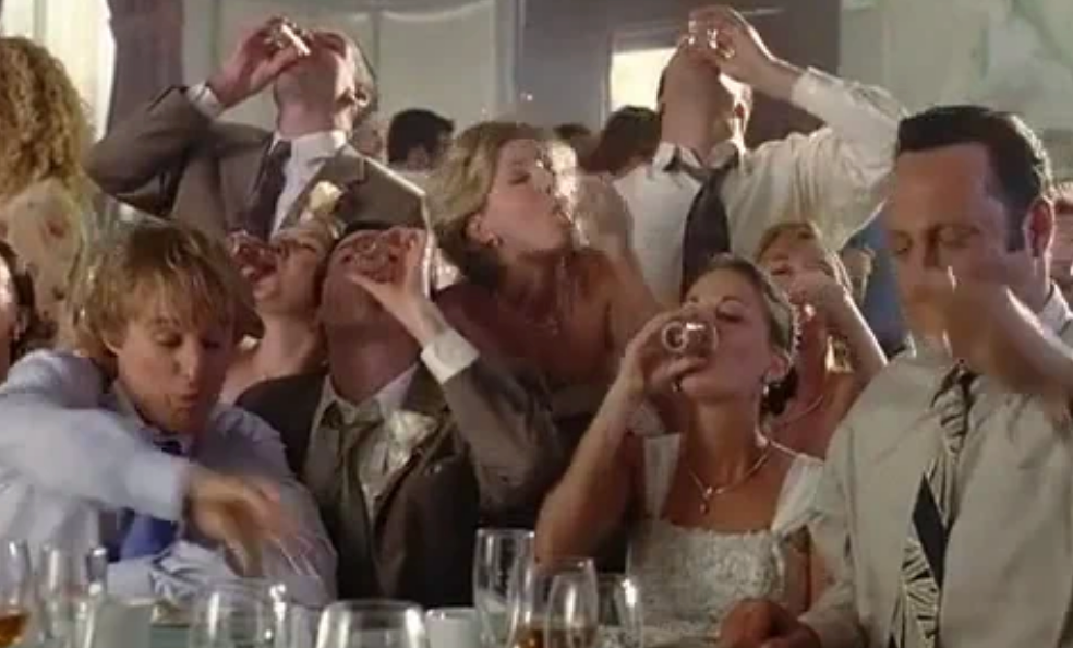 A group of people at a party, including a bride and groom, are seated at a table taking shots together