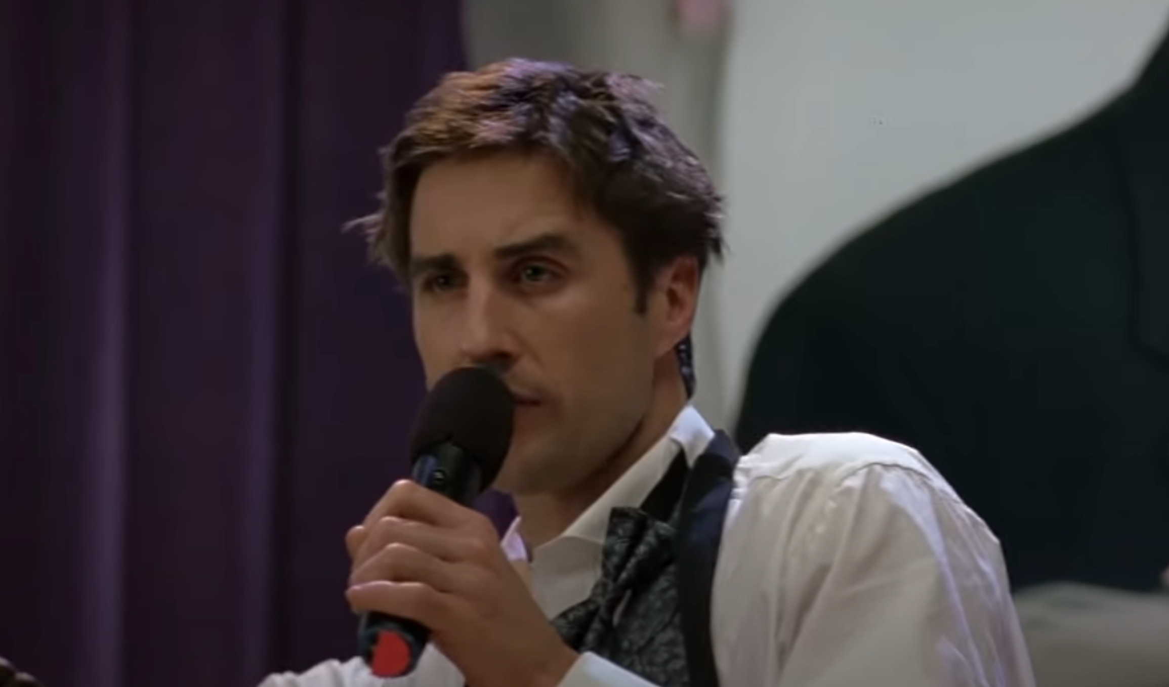 A person speaks into a microphone, wearing a light dress shirt and a loosened tie, in what appears to be a public speaking setting