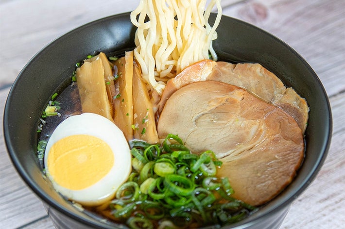 ラーメンの丼、具材は茹で卵、チャーシュー、ネギ、メンマがあり、箸で持ち上げた麺が見える。