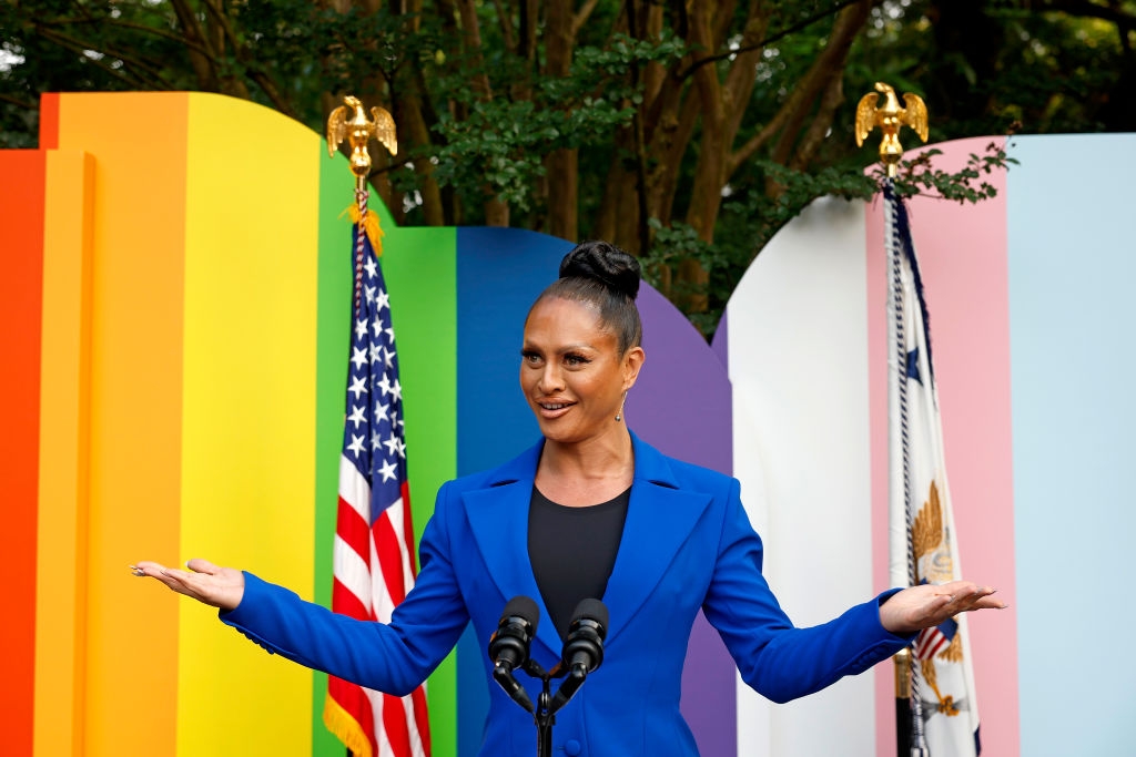 Person in a blue blazer speaks at a podium with microphones, standing in front of colorful panels and American flags