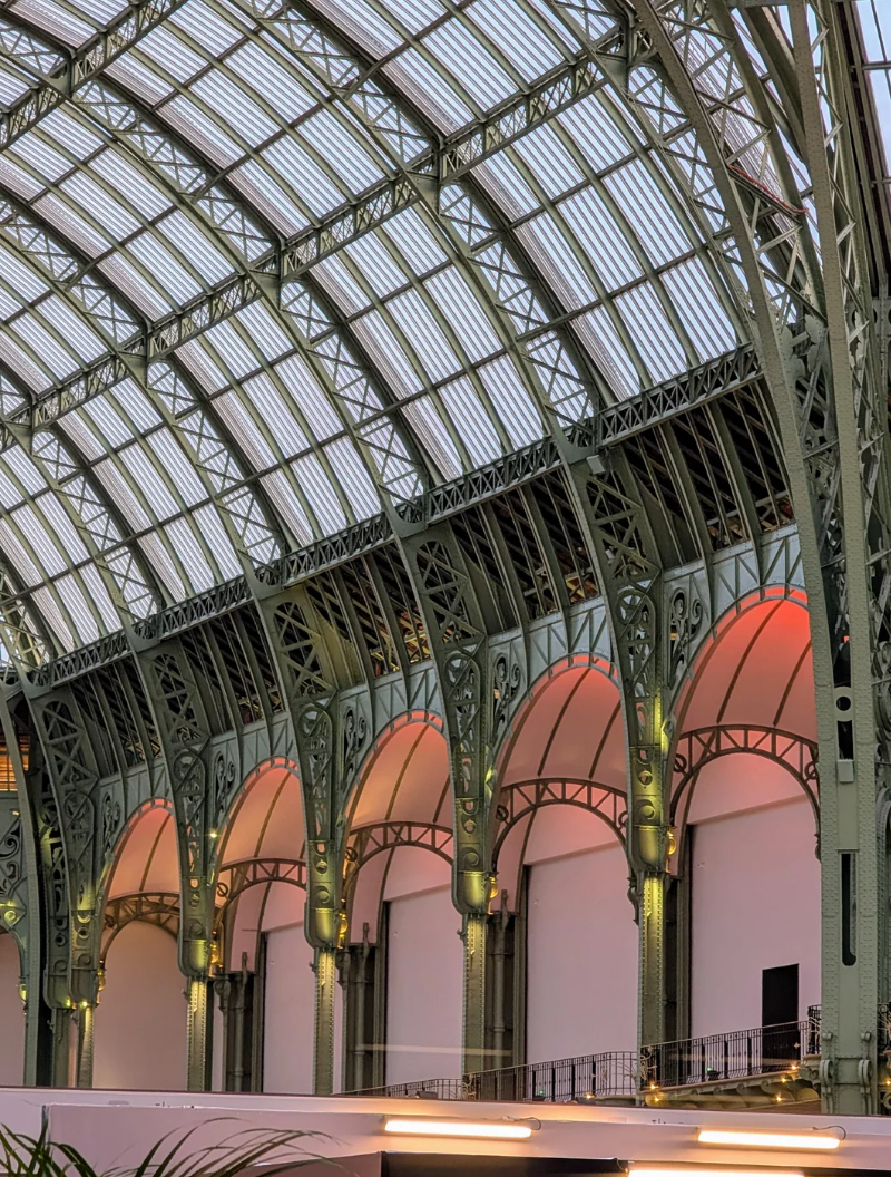 Interior view of a spacious hall with an ornate, arched glass ceiling and detailed metal framework. Soft lighting highlights architectural features