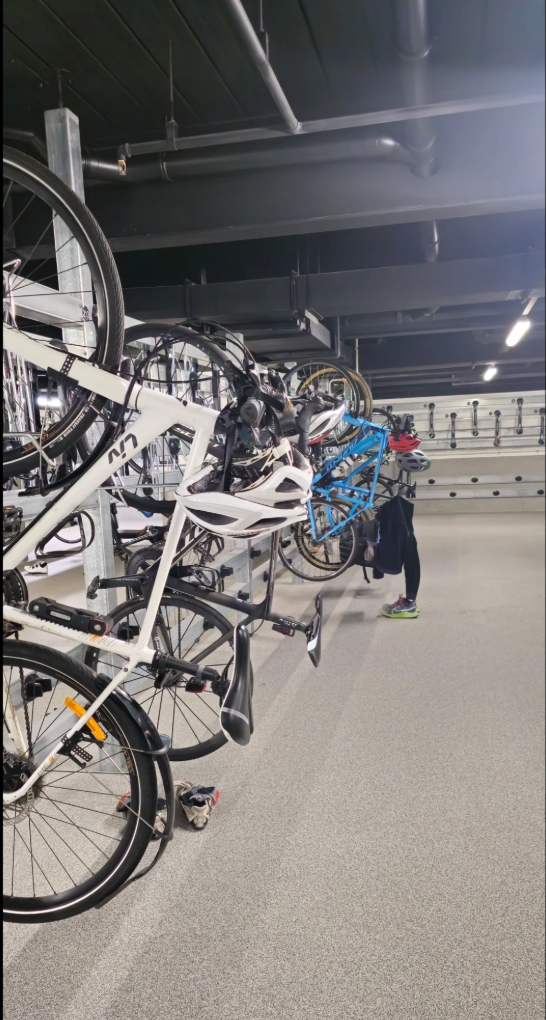 Bicycles are secured on a vertical rack in a parking area. A person in activewear adjusts a bike, and another bike is partially visible on the right
