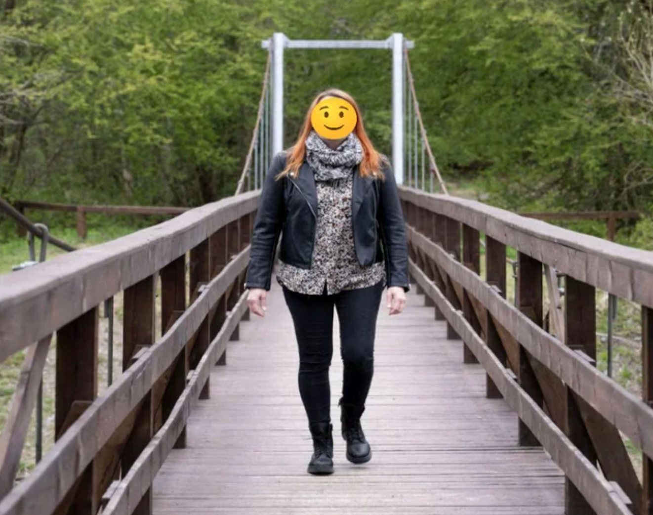 Person with a smiling emoji face stands on a wooden bridge in a jacket and patterned top, surrounded by a forest