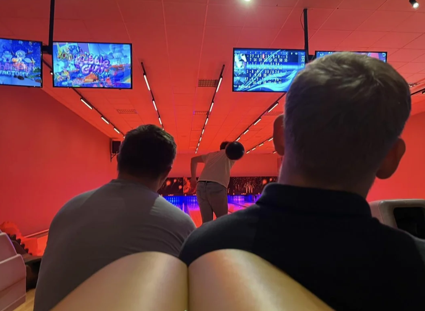 People watching a person bowl in a bowling alley with score screens above the lanes