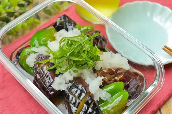 ガラス容器に盛られた焼きナスとピーマンの料理。大根おろしと刻んだシソが上にのっている。横に飲み物と皿がある。