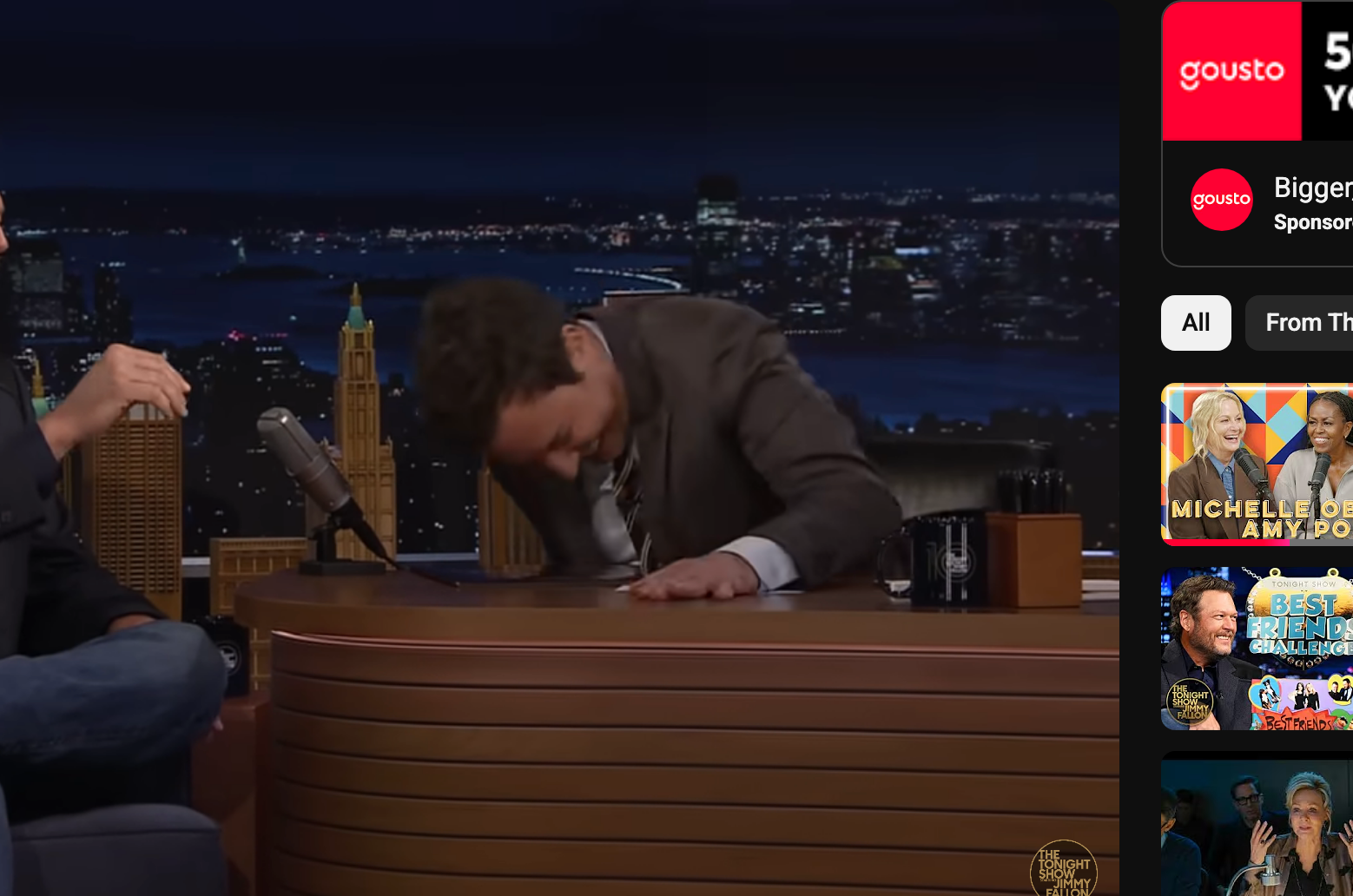 Jimmy Fallon laughing at a desk during a conversation with Blake Shelton on his talk show