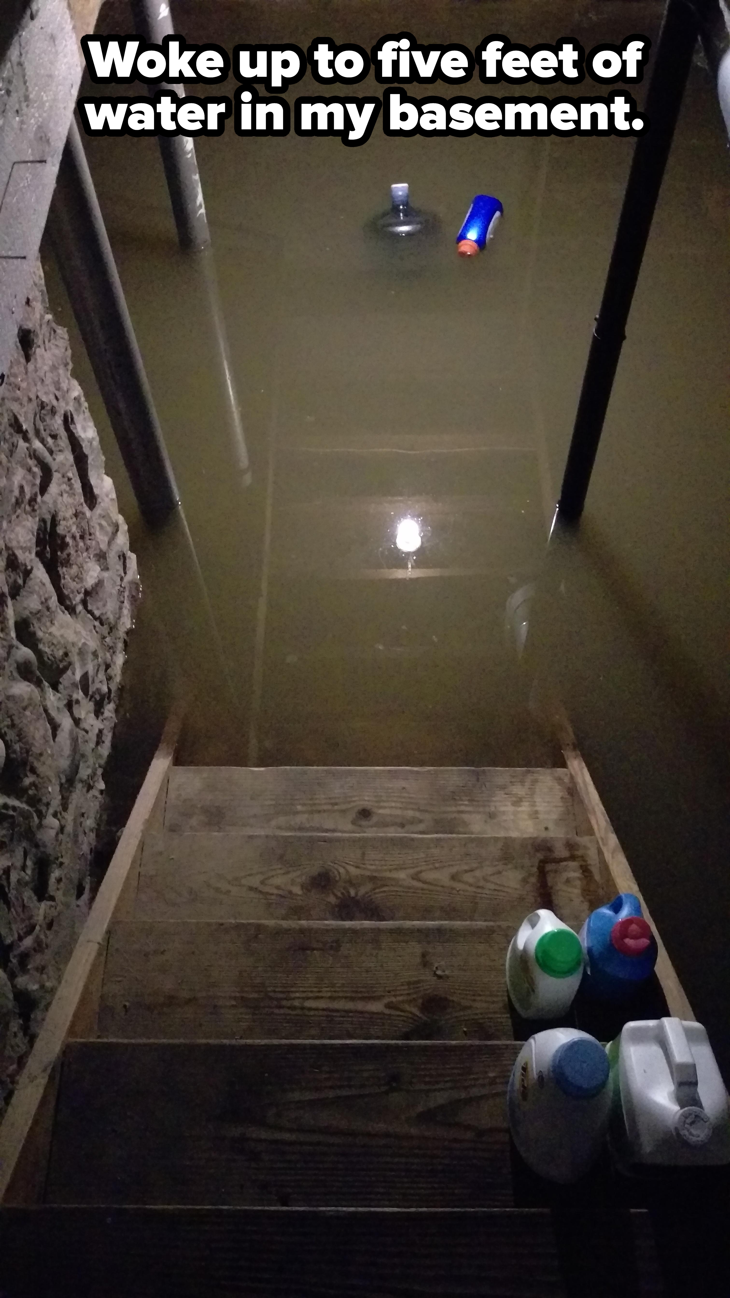 Flooded basement stairs with floating detergent bottles and submerged light reflecting on the water surface. Stone wall visible on the left