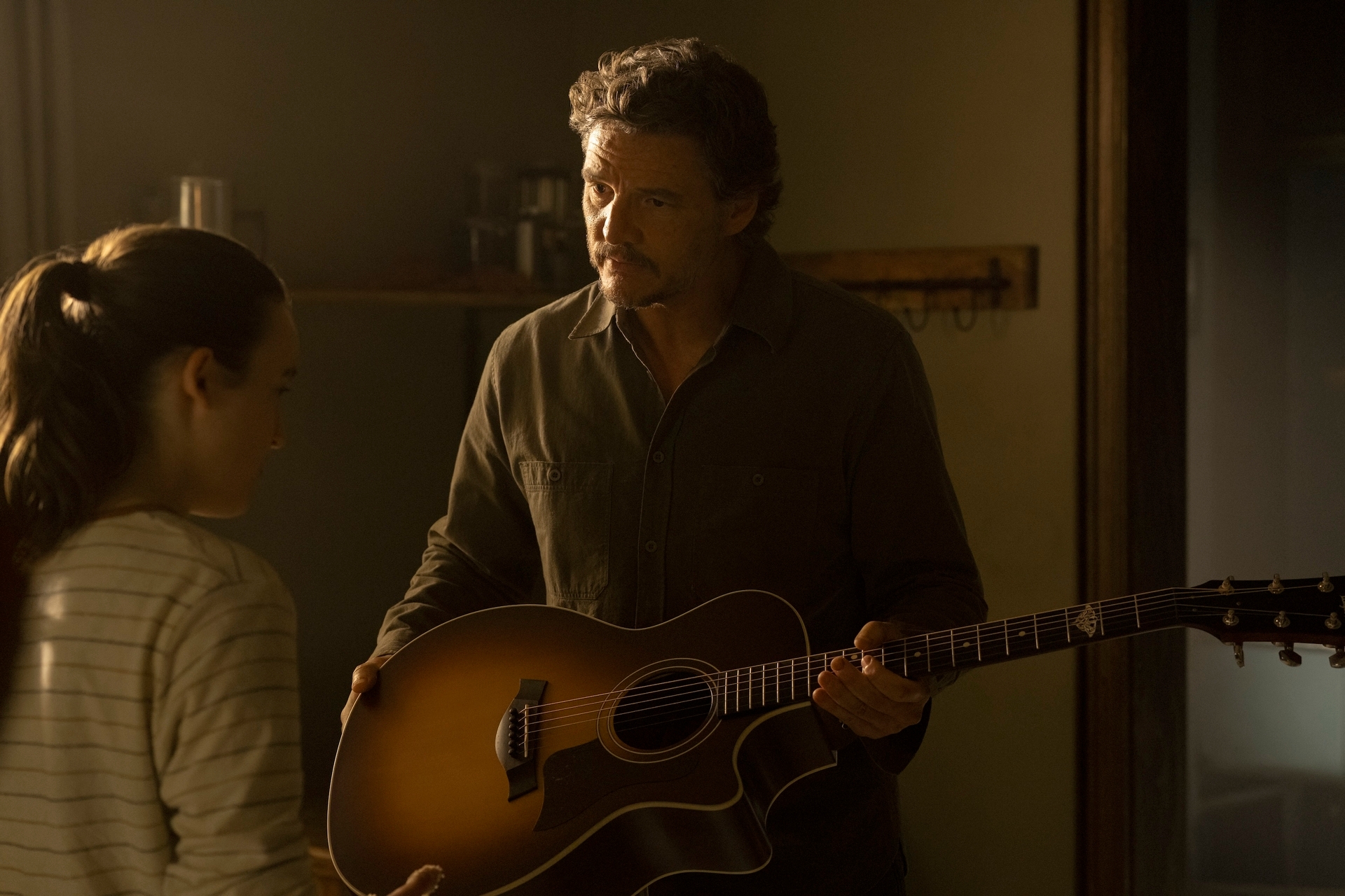 Hombre sosteniendo una guitarra mientras habla con una joven con camiseta de rayas. Ambiente informal en una habitación iluminada suavemente