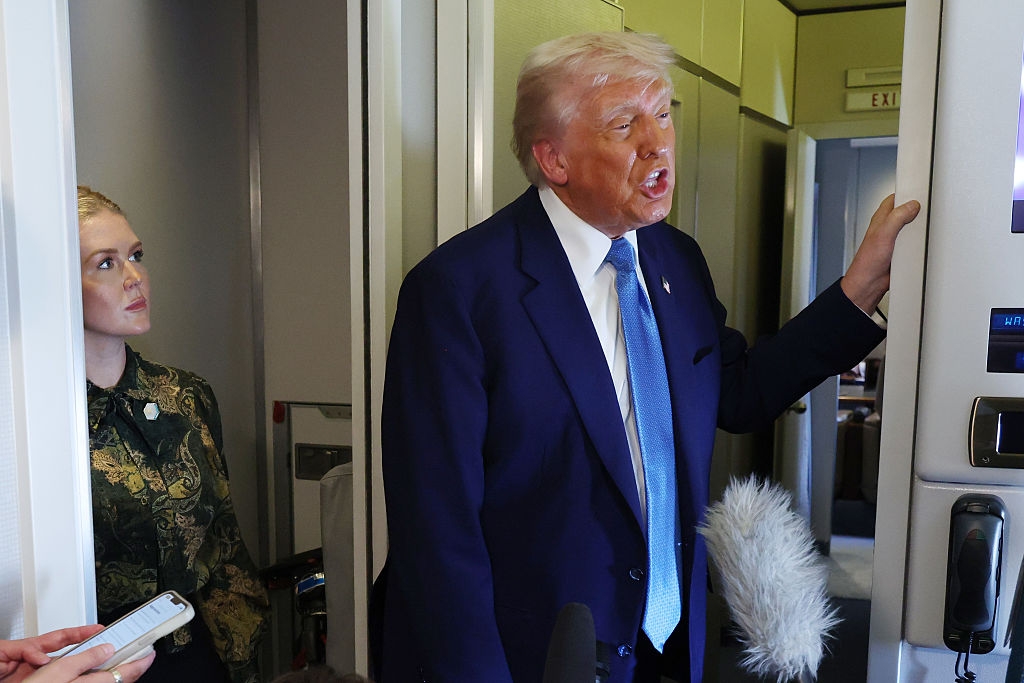 A person in formal attire speaks to reporters inside an aircraft, with a woman with long hair observing