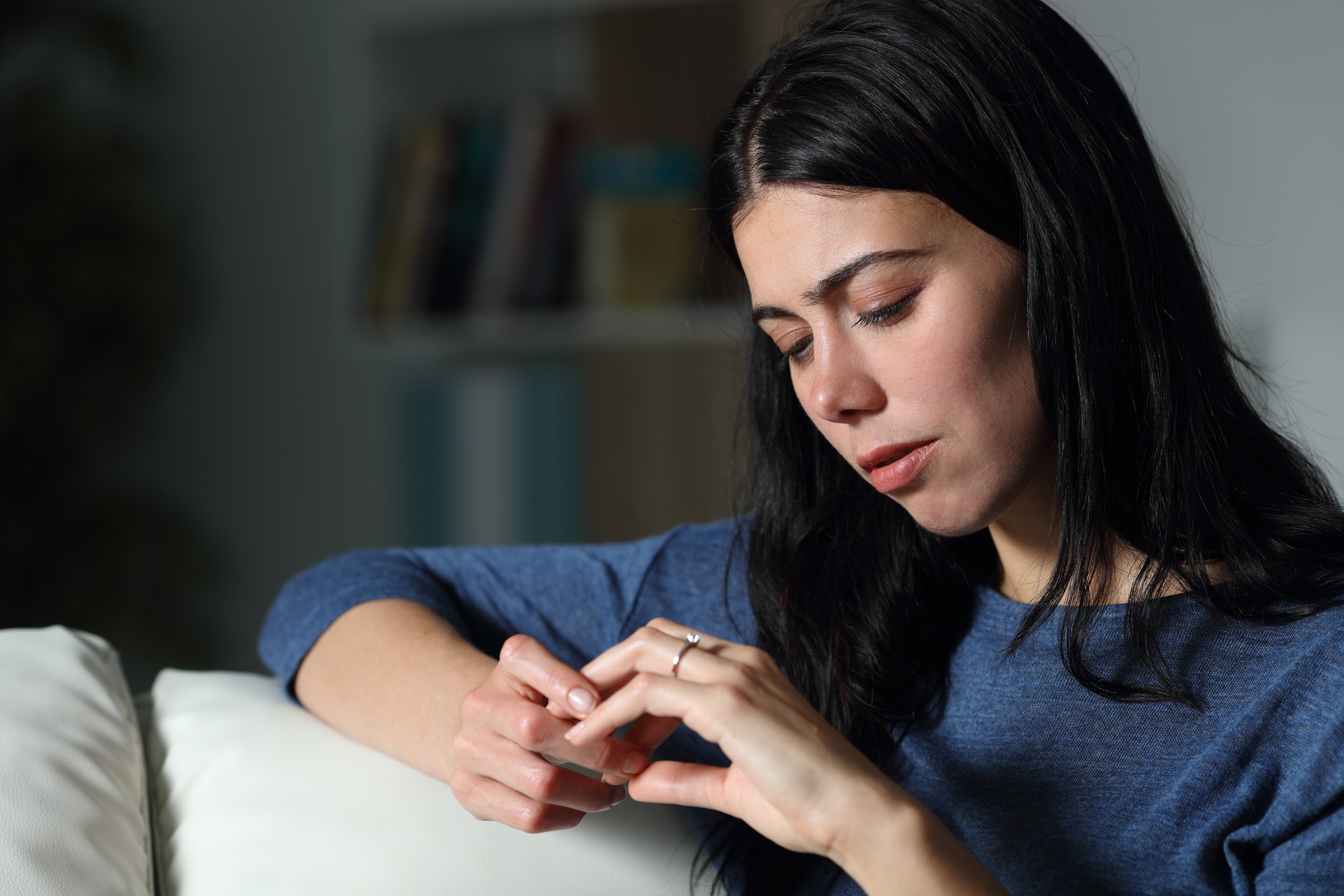A person sitting on a couch, looking thoughtful and touching their fingertips together, wearing a simple long-sleeved top