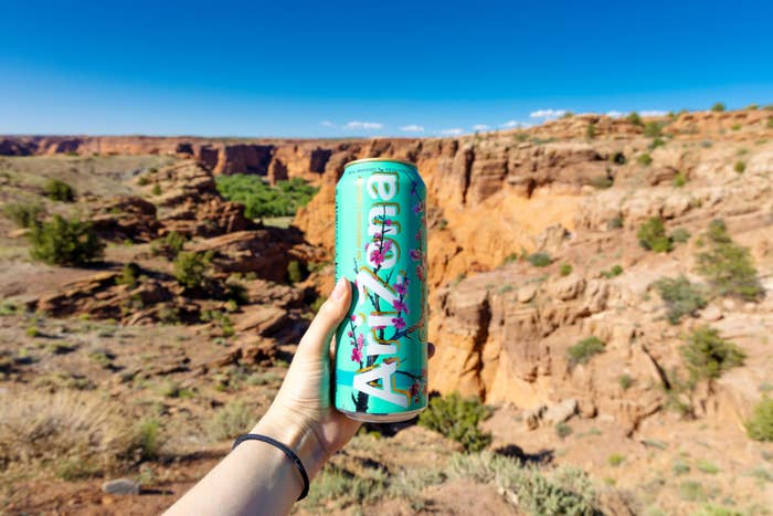 Person holding a can of Arizona Tea in front of a rocky canyon landscape under a clear sky