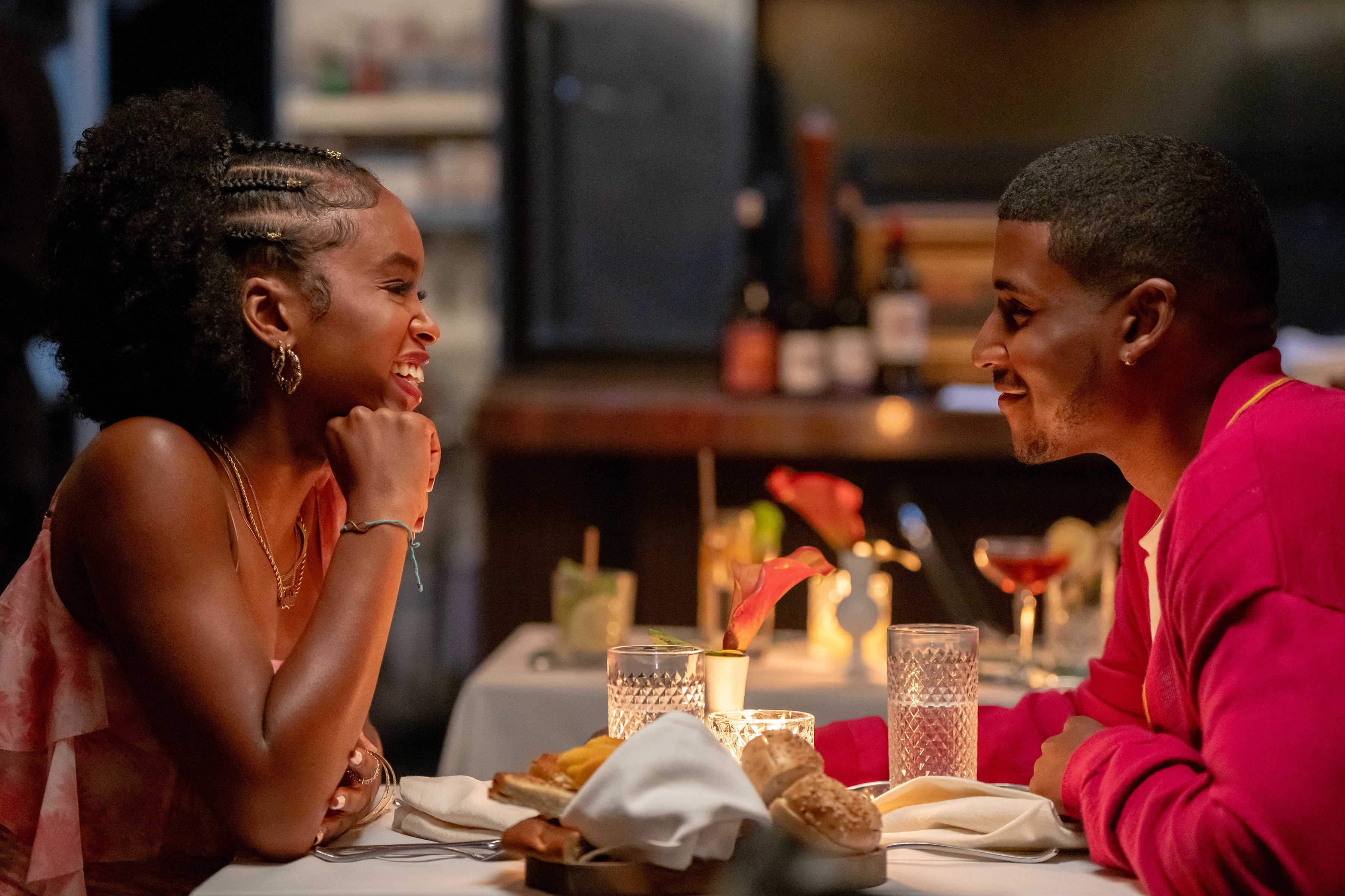 Two people sitting at a candlelit dinner, smiling and engaged in conversation. The setting appears romantic and cozy