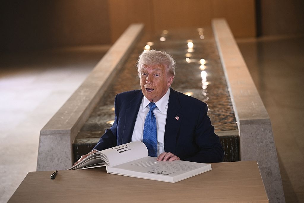 Person in a suit looking at a large open book on a table, seated near a reflective water feature indoors