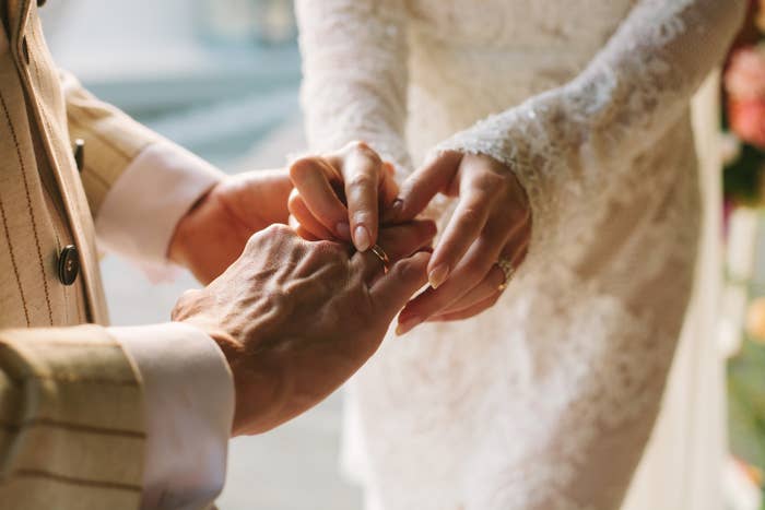 A person places a ring on another's finger during a wedding ceremony. Both wear formal attire, with the focus on hands and the symbolic exchange