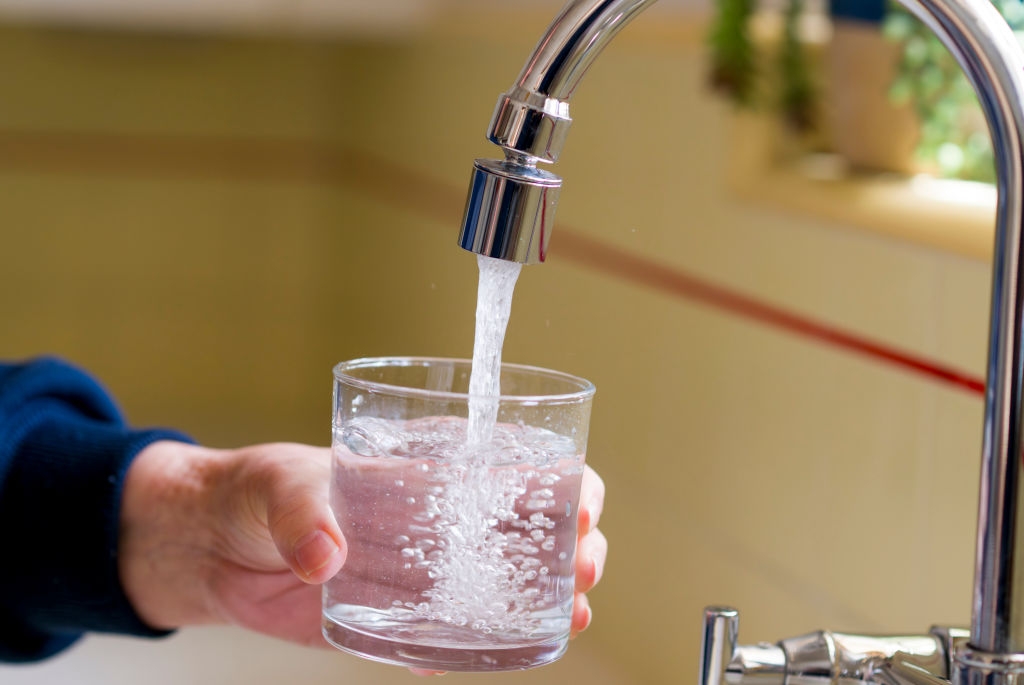 A person fills a glass with water from a kitchen faucet, focusing on the stream entering the glass