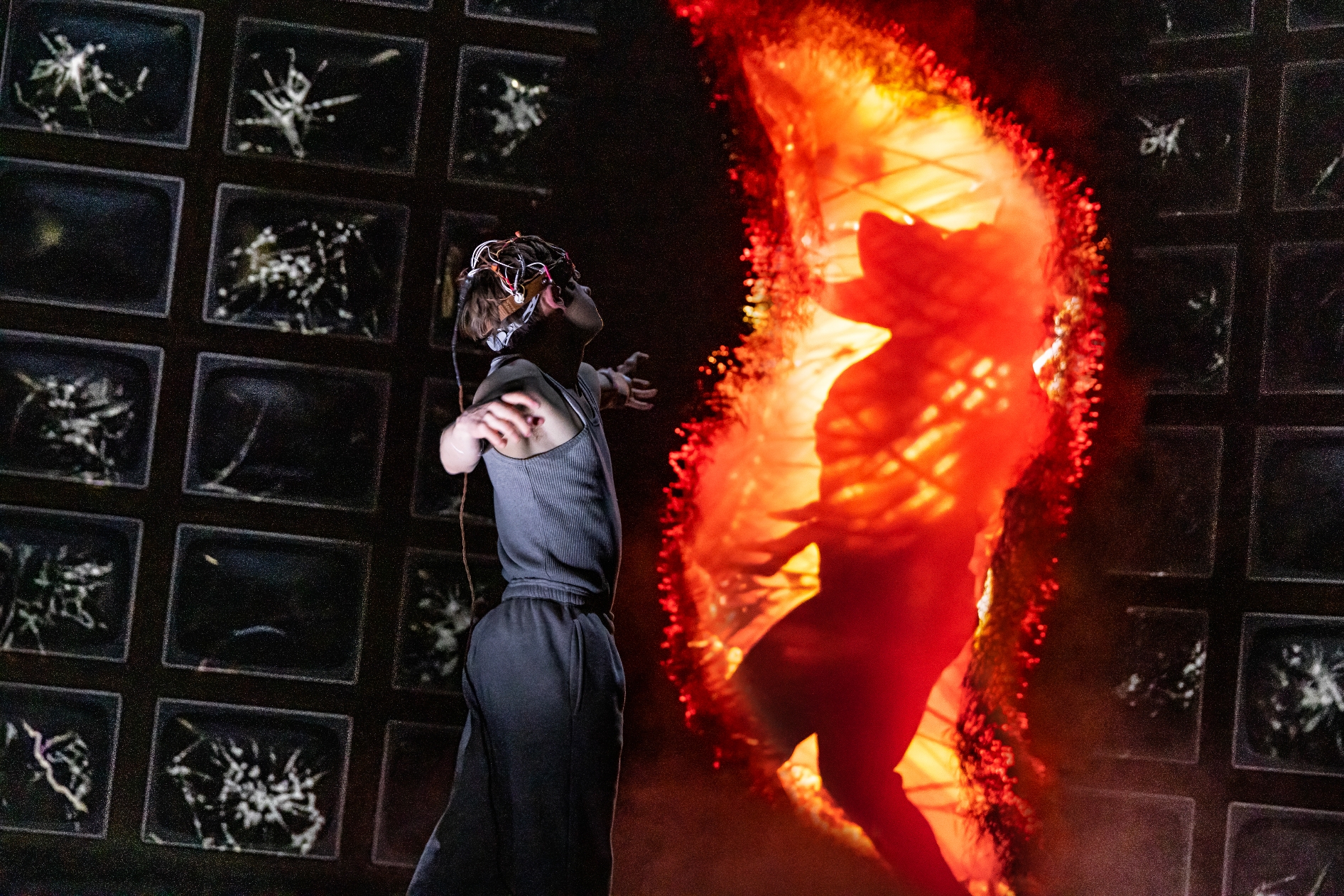 A performer, wearing a headpiece, poses dramatically in front of a fiery silhouette backdrop on stage