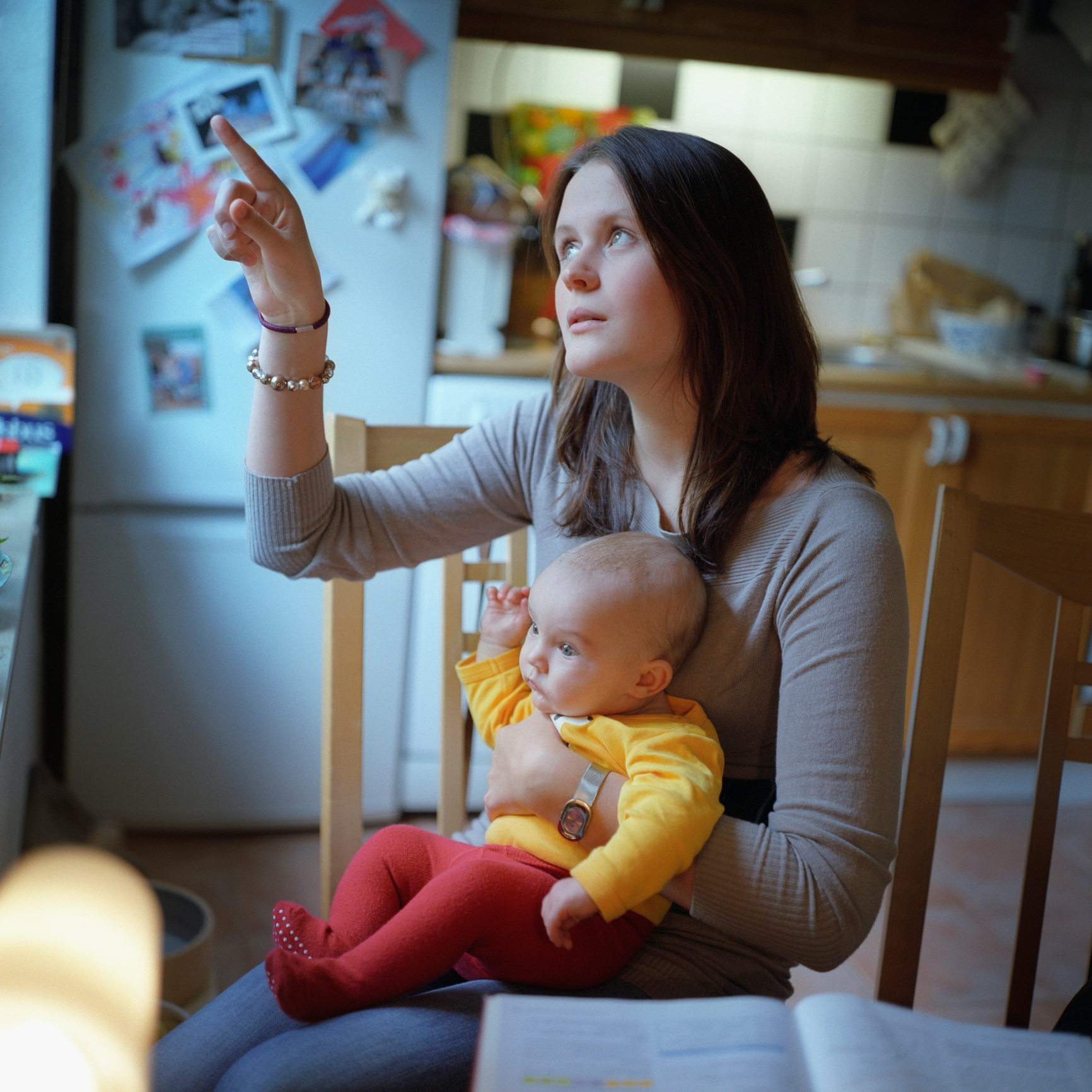A woman holds a baby on her lap in a kitchen, gesturing and looking upward thoughtfully