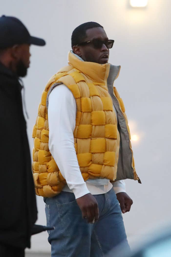 Man in quilted vest, white long-sleeve shirt, and jeans, walking outside with another person