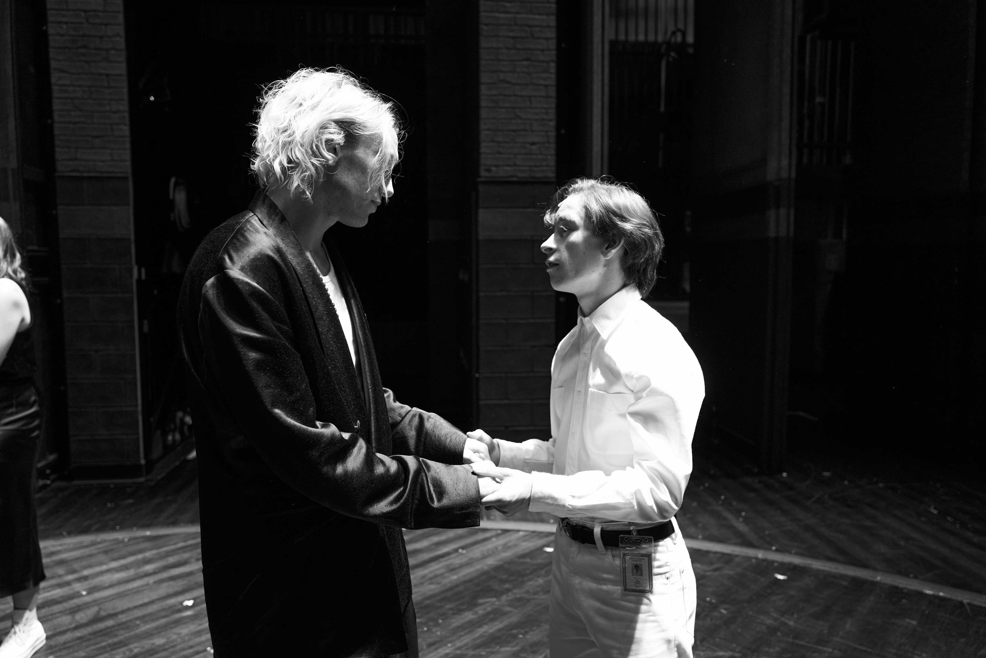 Two individuals stand facing each other, holding hands, in an emotional moment backstage. One wears a formal suit, the other a white outfit