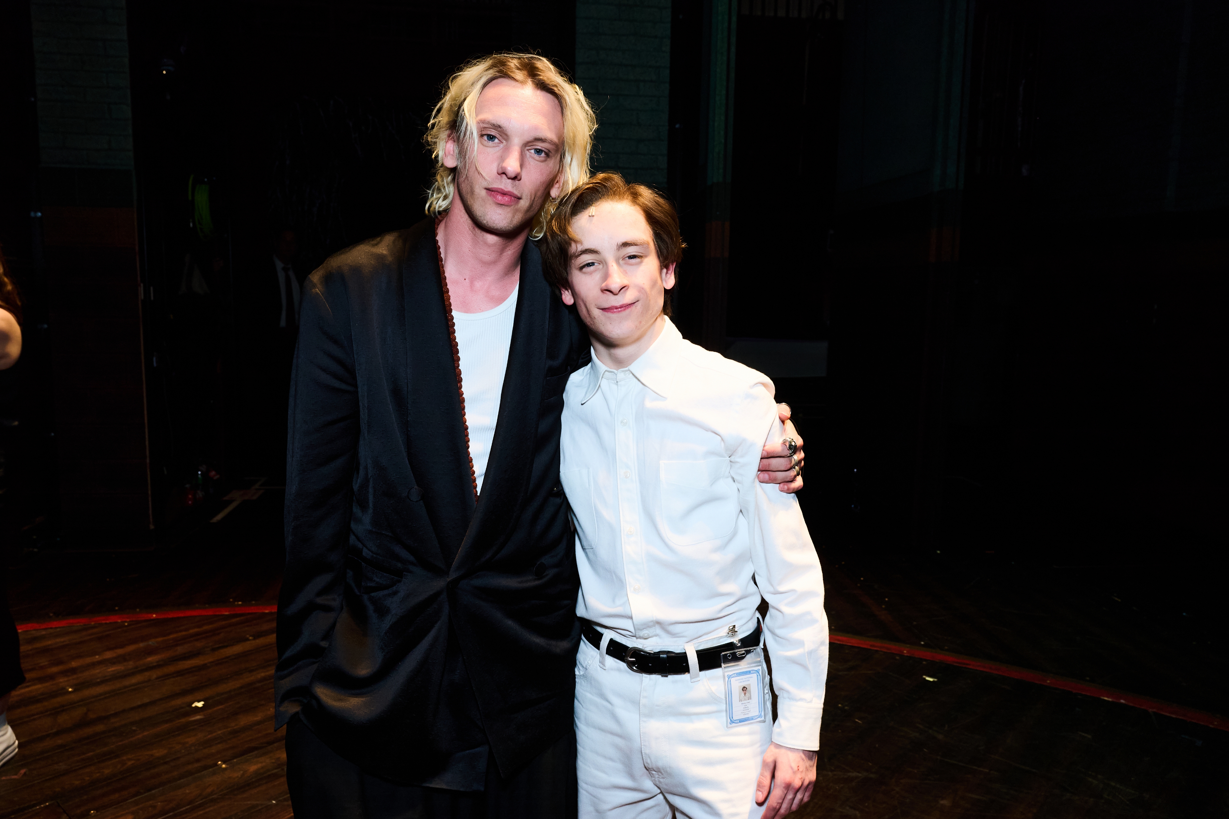 Two individuals pose together at an event; one wears a black coat, the other wears an all-white suit with an ID badge