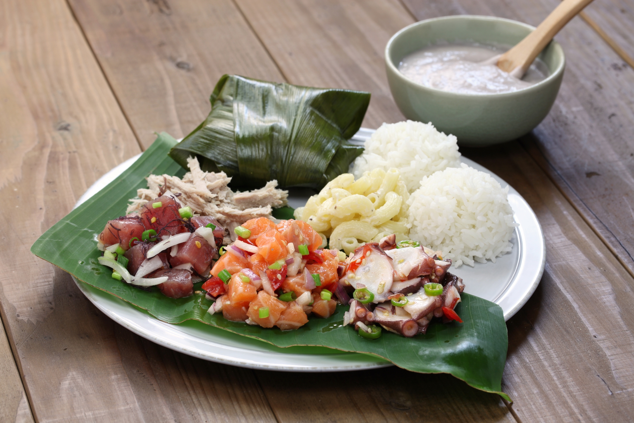 Plate with a variety of Hawaiian dishes: poke, macaroni salad, rice, and a bowl of soup on a wooden table