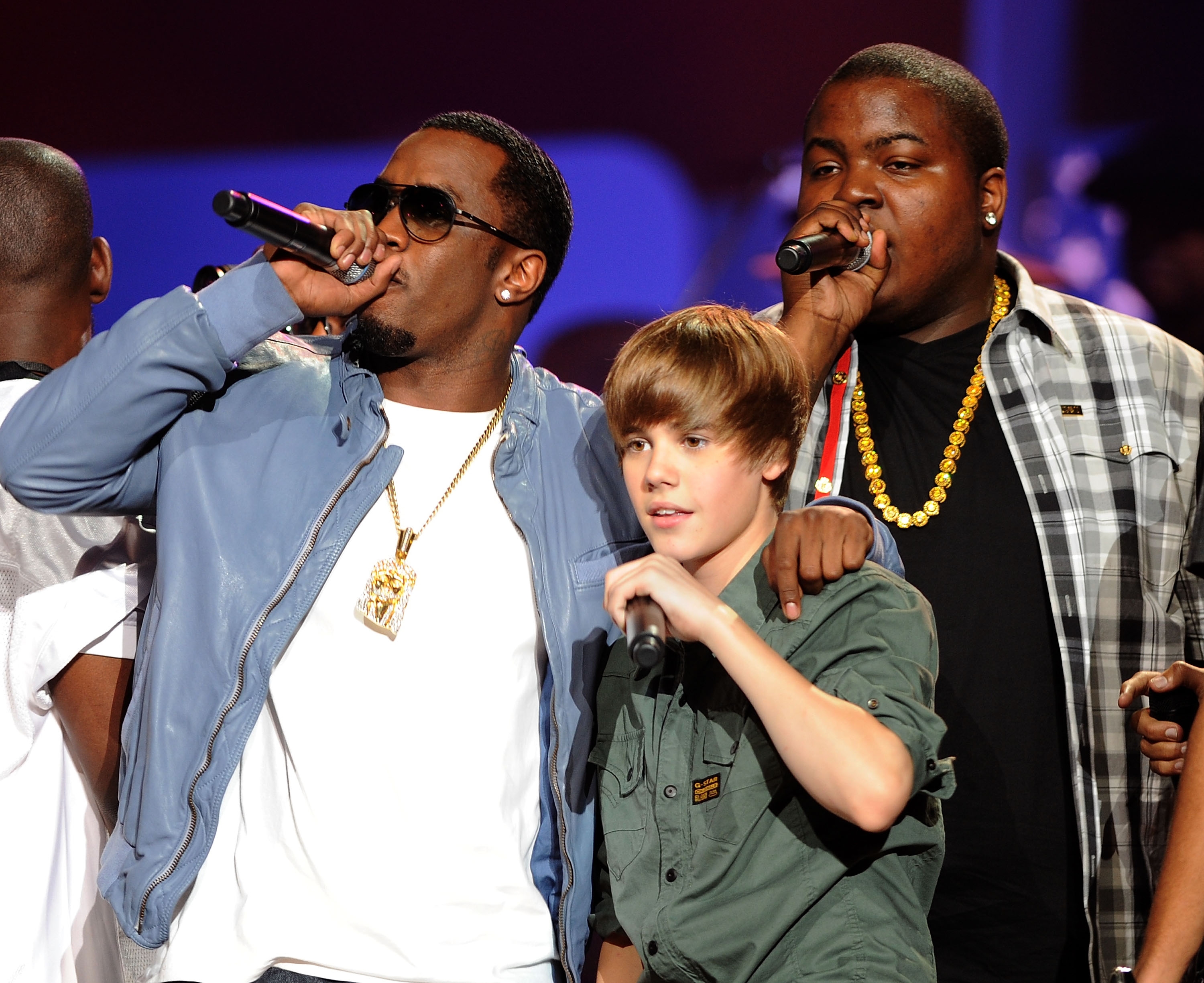 Three performers on stage, singing into microphones. The center person is wearing a casual shirt, flanked by two with bold accessories and jackets