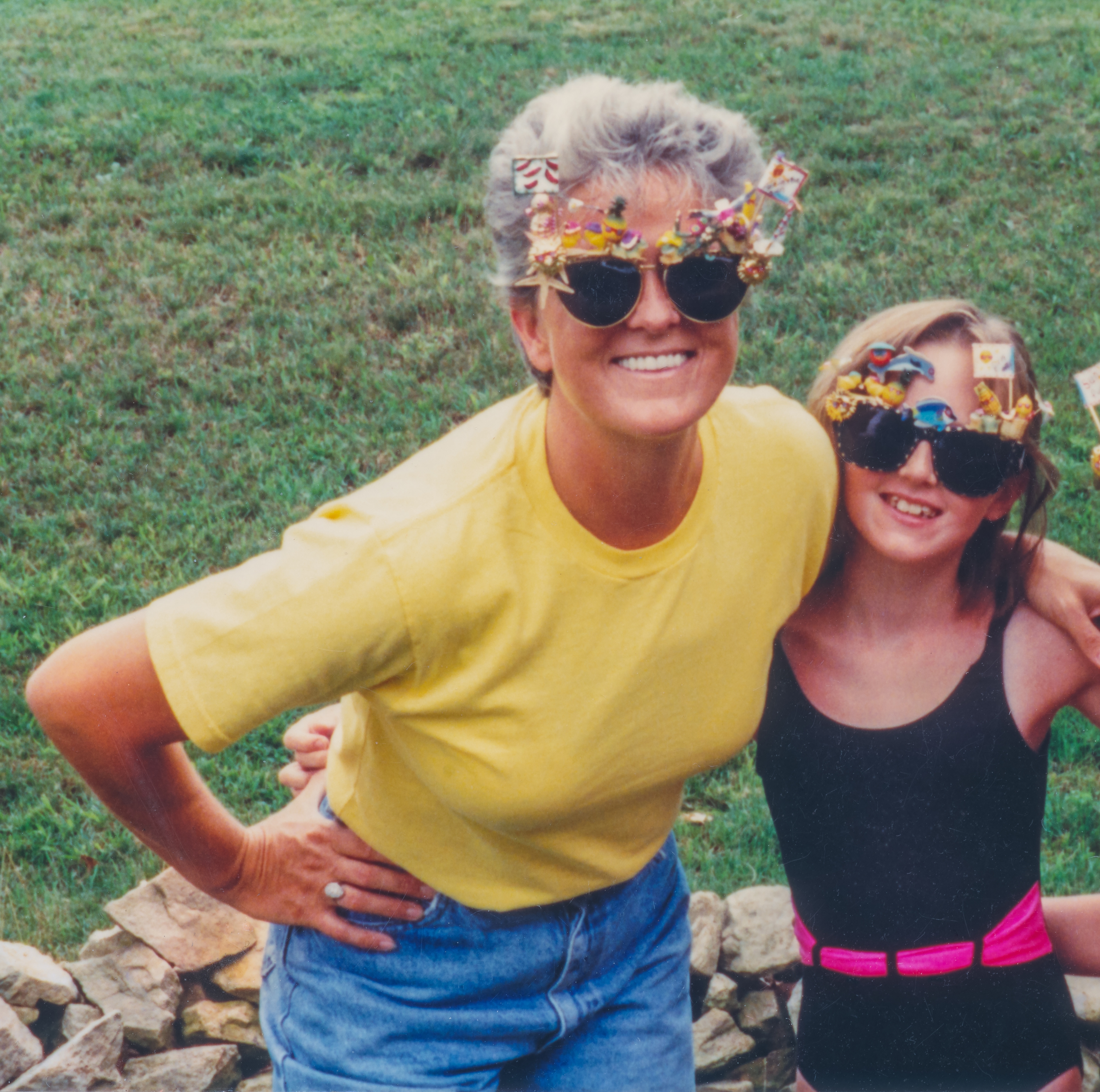 Two women outdoors, both wearing playful decorated sunglasses and smiling. They appear to enjoy a moment together in a grassy yard
