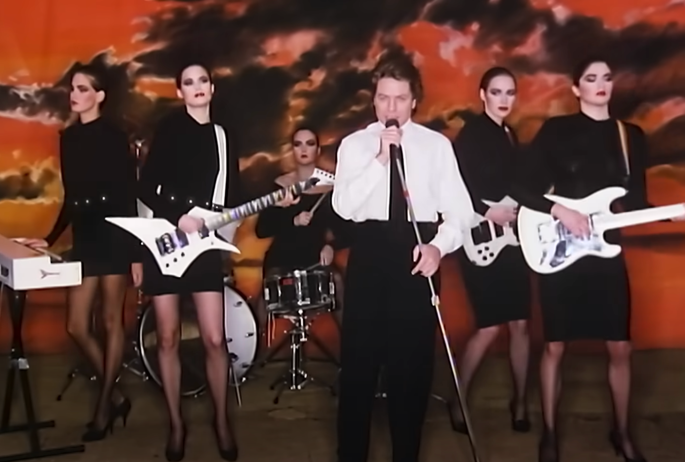 A band performs against a painted backdrop. The lead singer holds a mic, wearing a suit. Four women in stylish black outfits play instruments