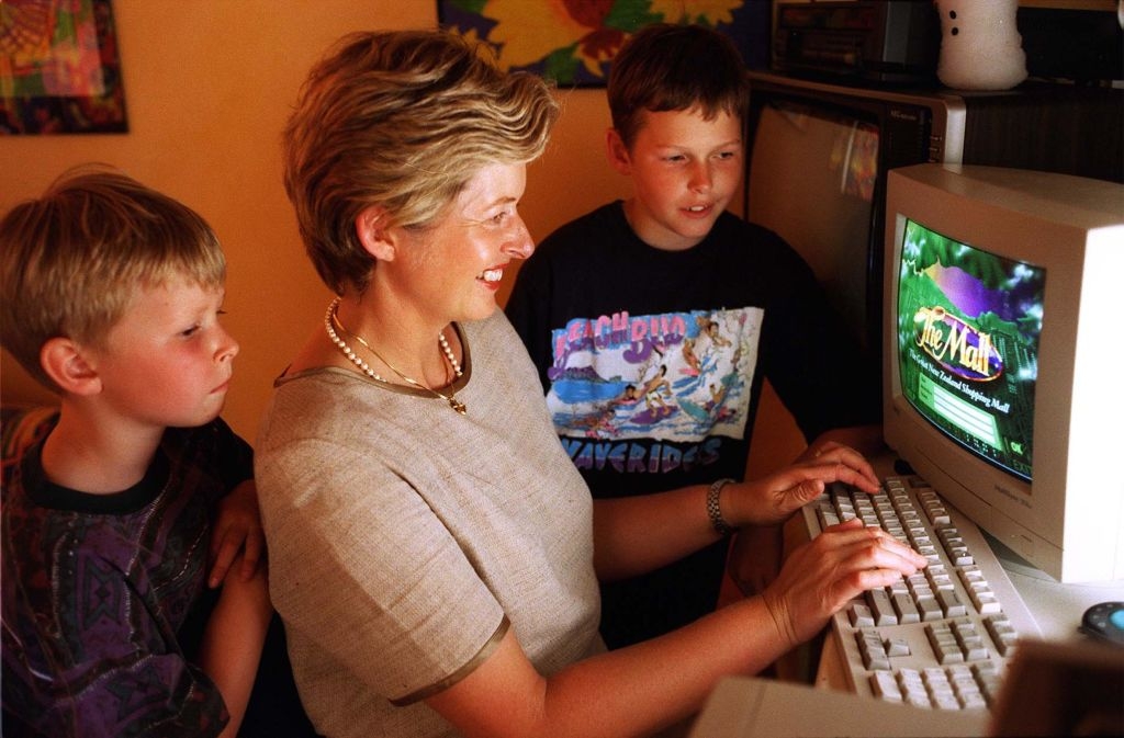 A woman sits at an old computer with two young boys watching, all engaged. The screen shows the game "The Magic School Bus Explores the Rainforest."