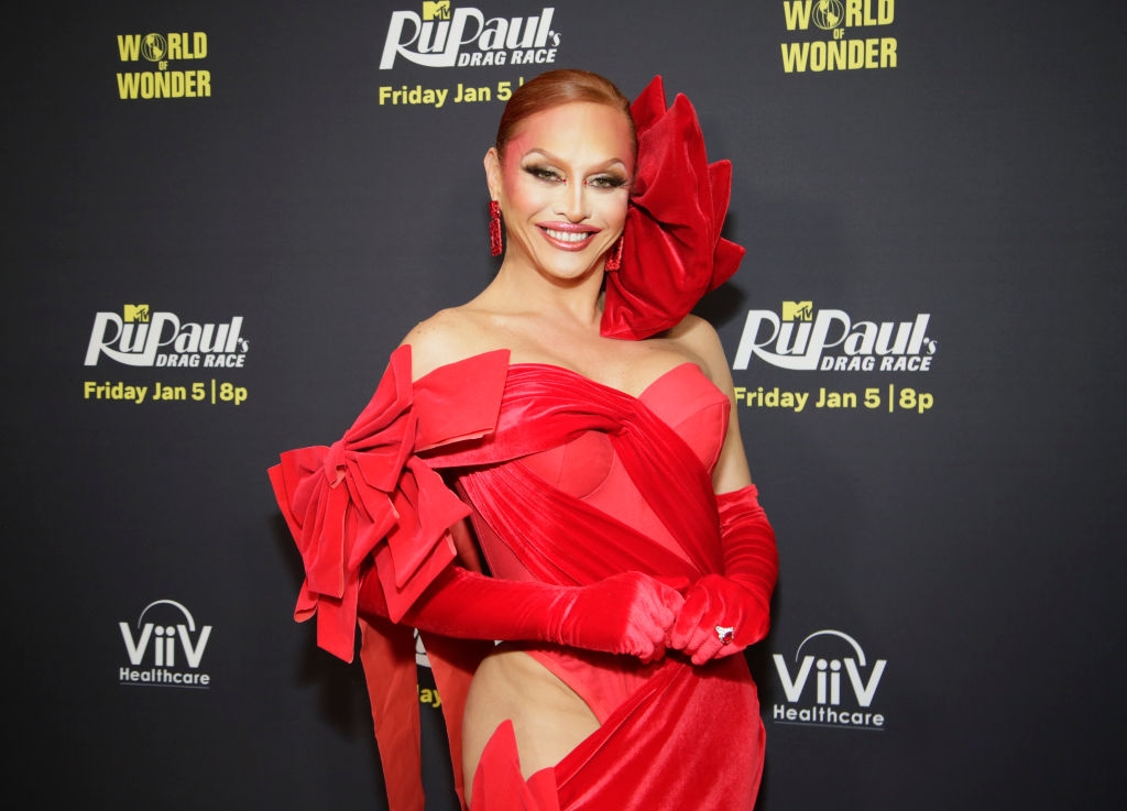 Person in an elaborate red gown with dramatic shoulder and hip details, smiling at a "RuPaul's Drag Race" event backdrop