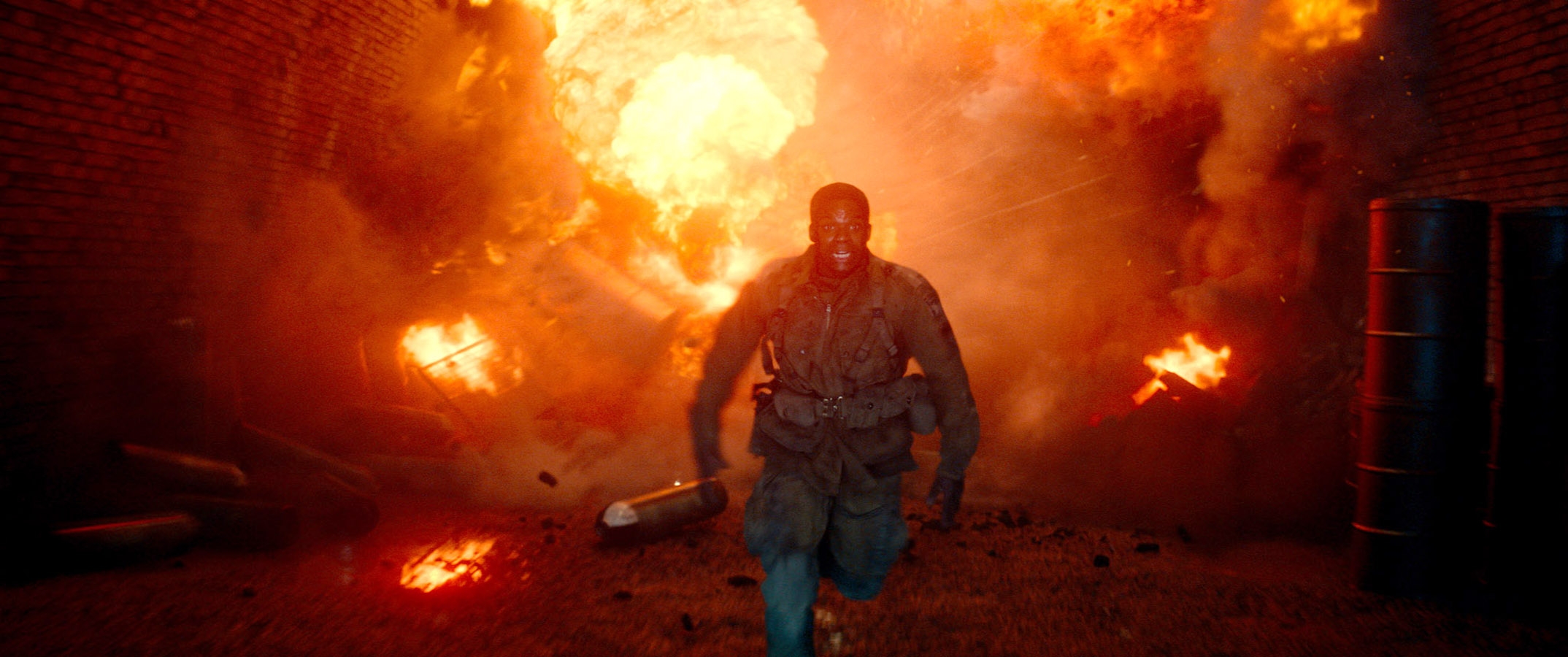 Boyce, wearing tactical gear, runs from a fiery explosion in an industrial setting
