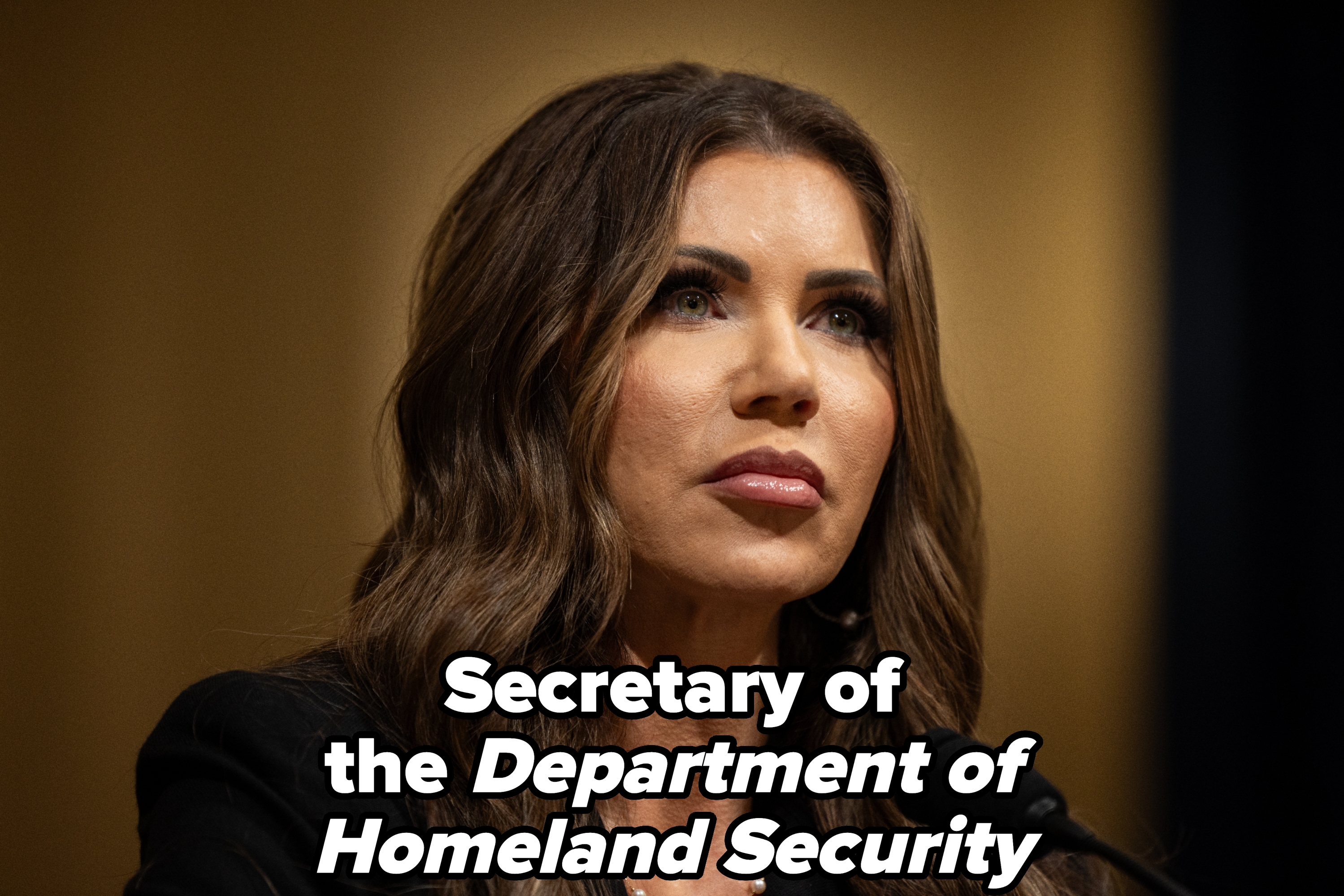 A person with long wavy hair speaks into a microphone at an event, wearing a formal dark outfit