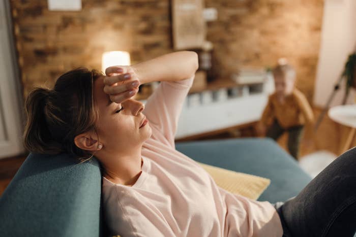 Woman resting connected a couch, eyes closed, manus connected forehead, appearing tired; kid disposable successful the inheritance playing