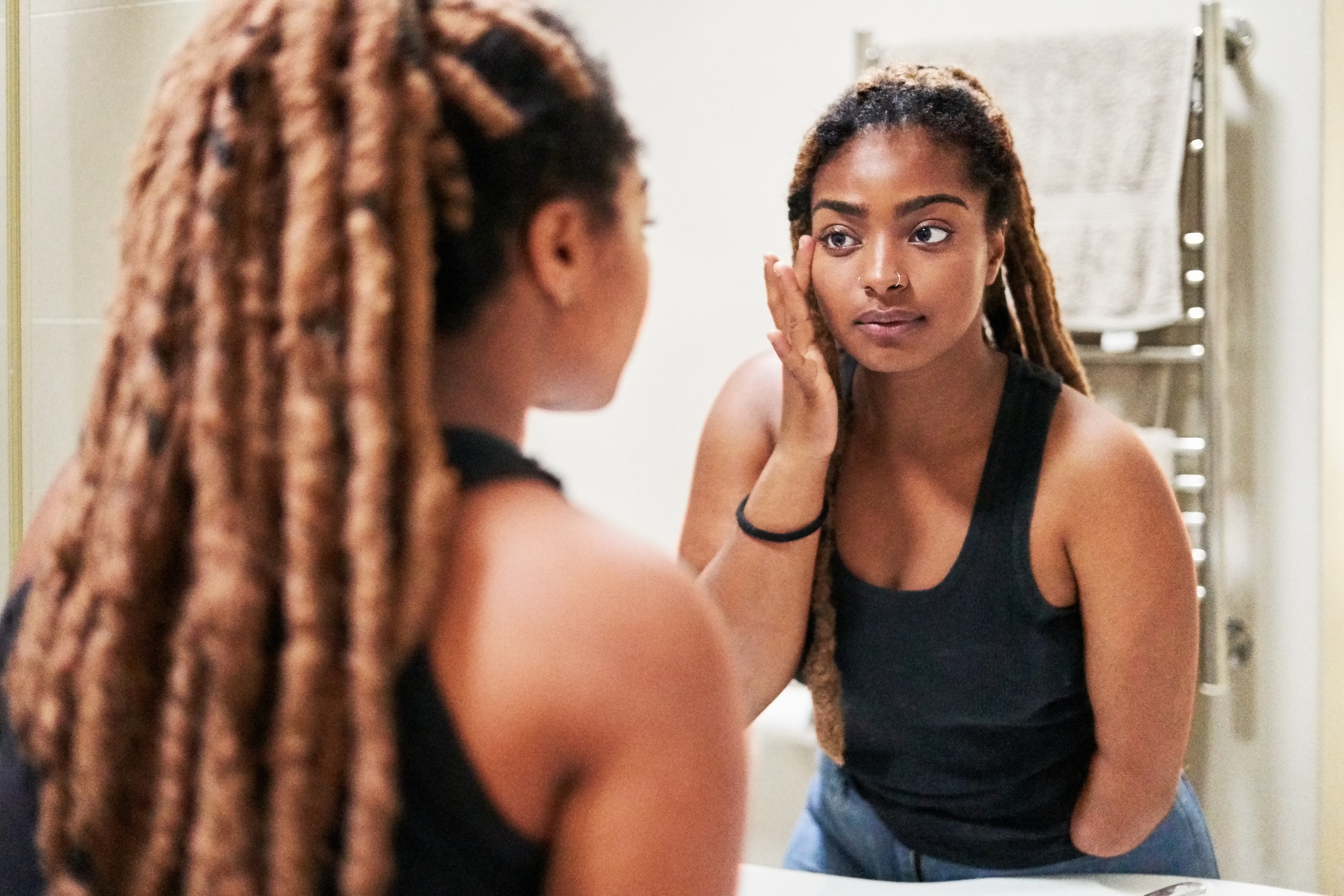 Woman with long braided hair, wearing a tank top, looks at her reflection in a bathroom mirror while touching her face