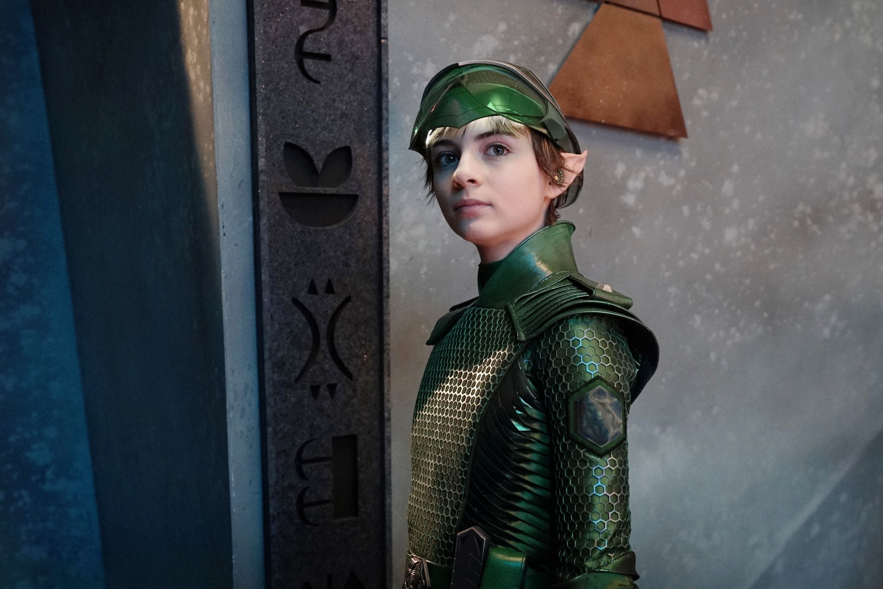 Person in a textured, futuristic costume with helmet and pointed ears, standing near symbols on a wall