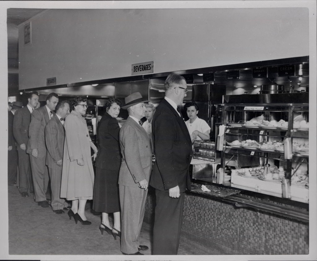 A line of people in 1950s-style attire wait at a cafeteria counter under a "Beverages" sign, showcasing mid-century dining culture