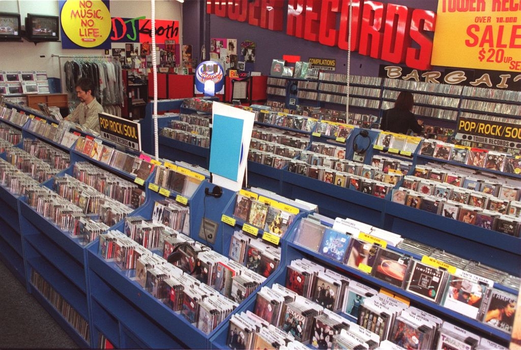 A bustling record store with rows of CDs and promotional signs, including one with "Tower Records" and sale notices. A shopper browses the aisles