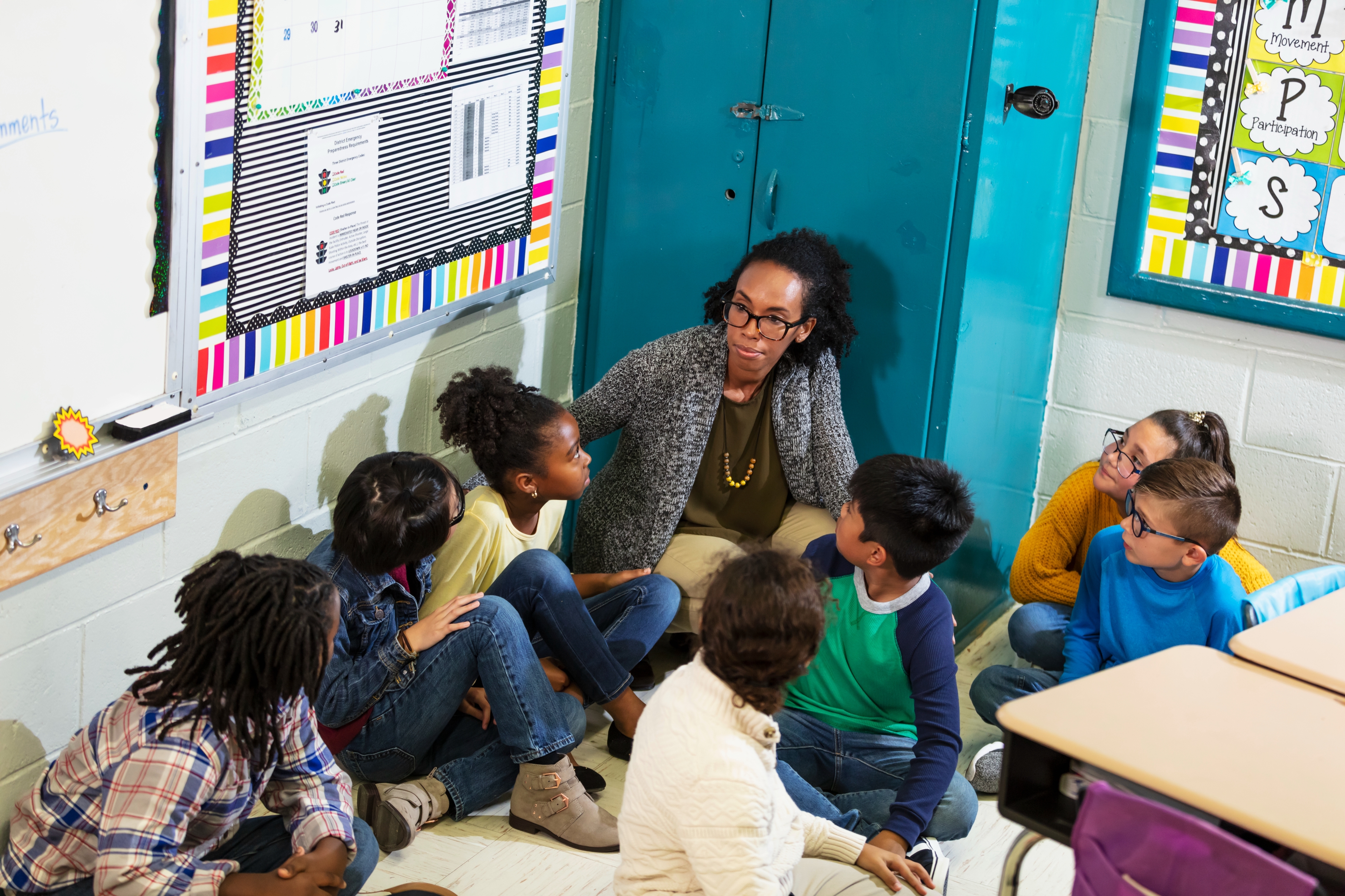 Teacher sitting on the floor with children in a classroom, engaging in discussion. The scene suggests an interactive learning activity