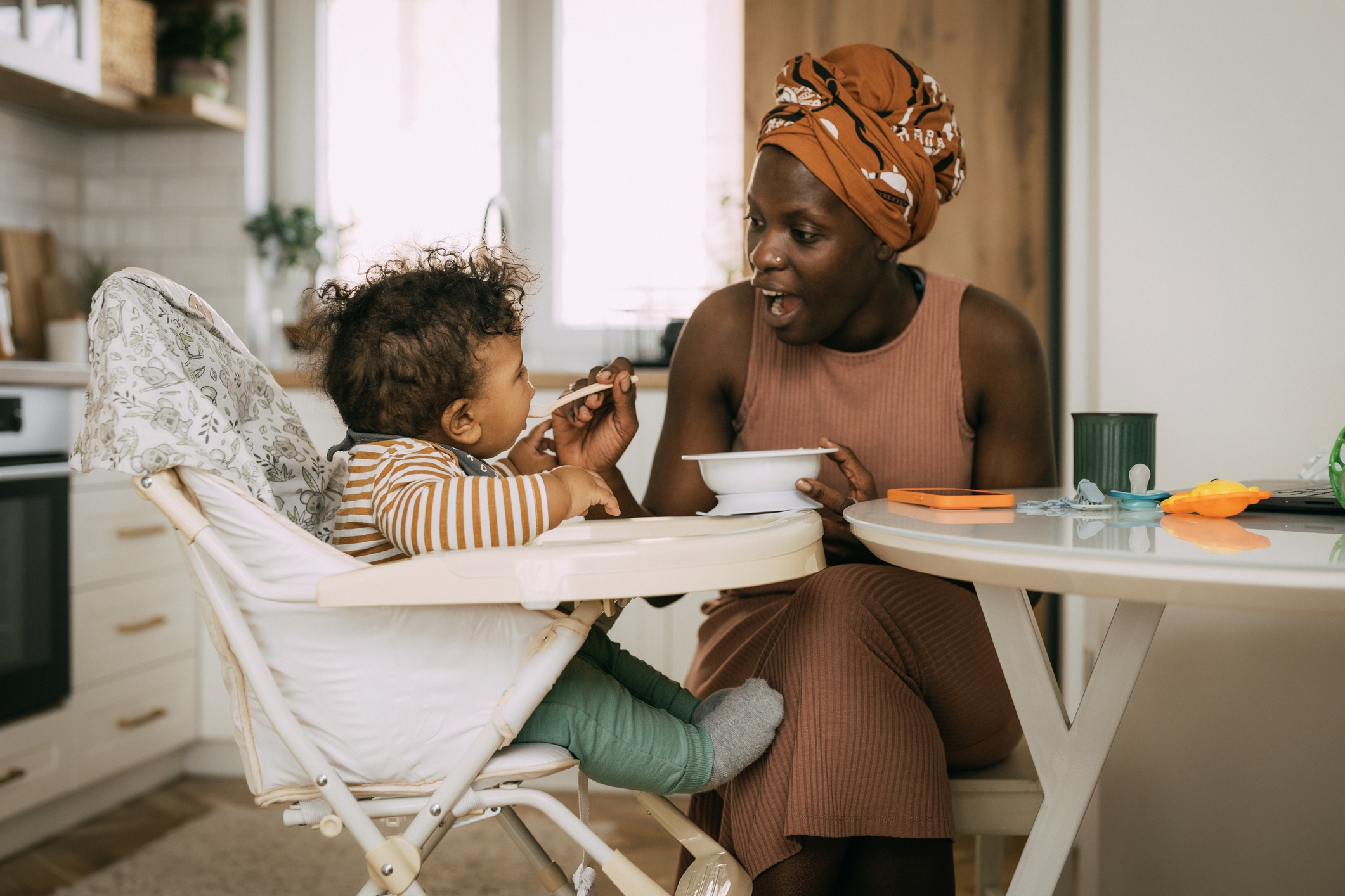 Parent feeding a smiling toddler in a high chair at a kitchen table, fostering a nurturing and joyful mealtime interaction