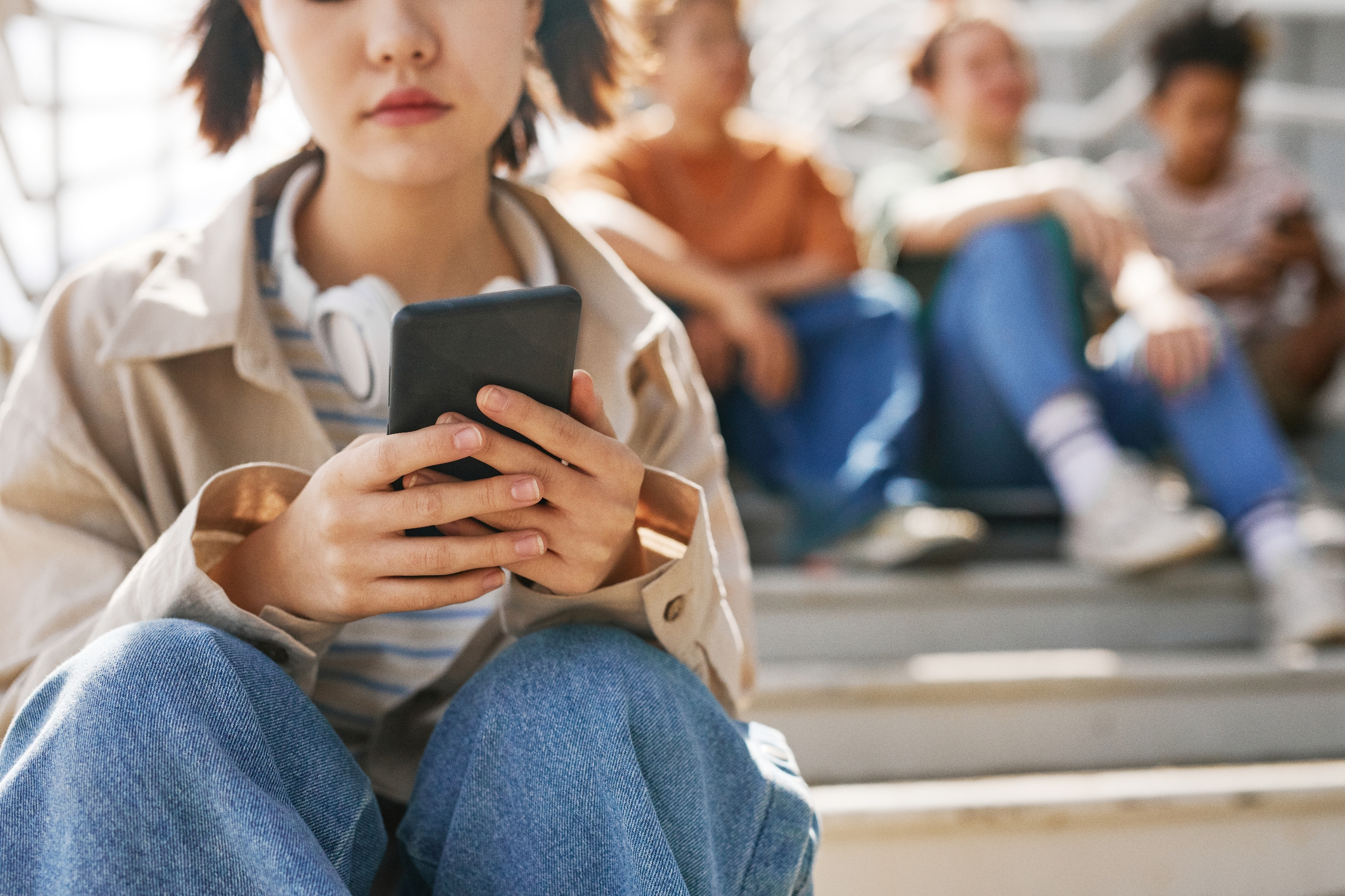 Teen sitting on steps, focused on phone, with friends chatting in the background. Casual attire. Context: teen social dynamics and technology use