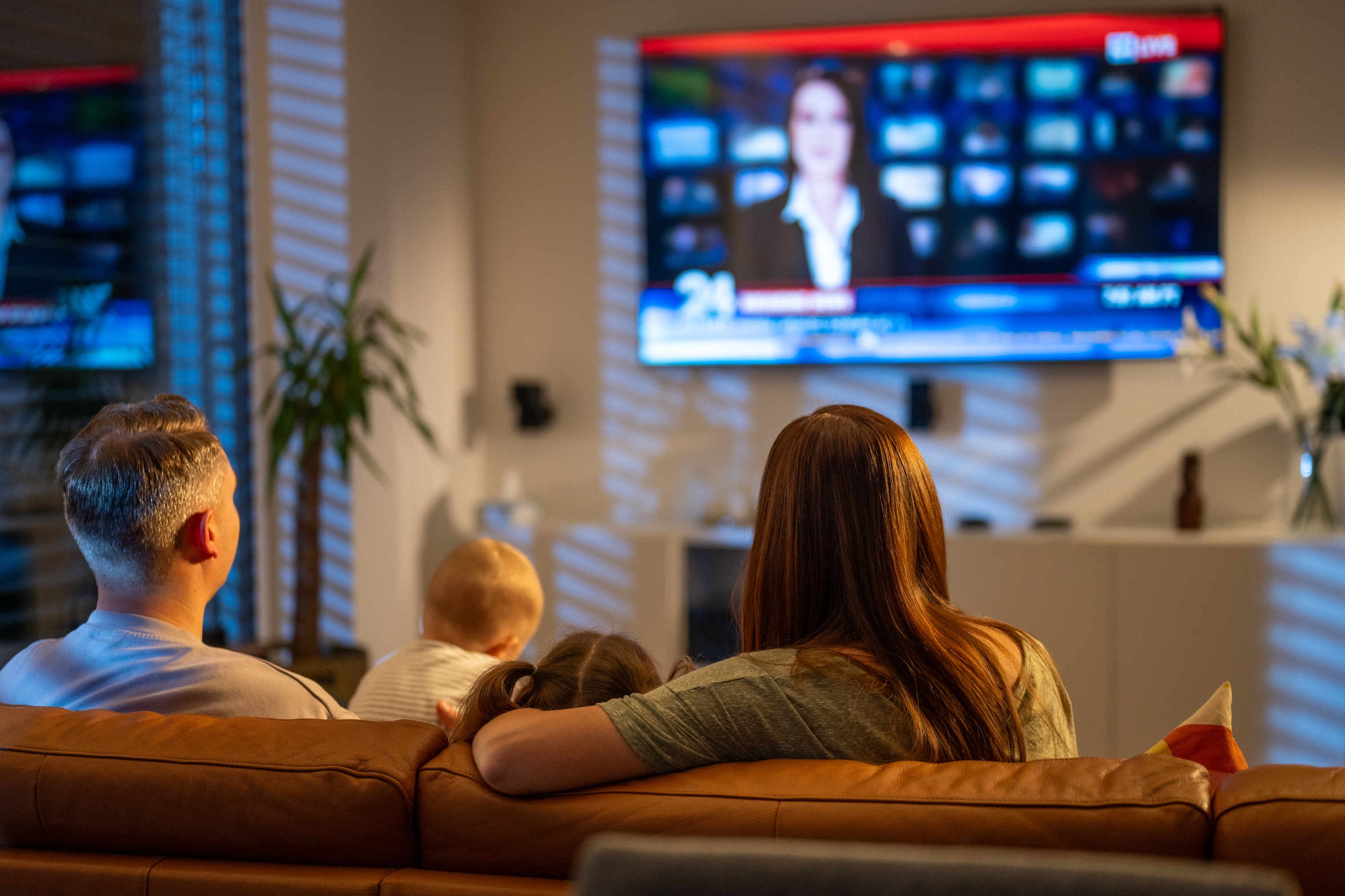 Family sitting on a couch watching a news program on TV; a baby is between two adults. Cozy home setting with soft lighting