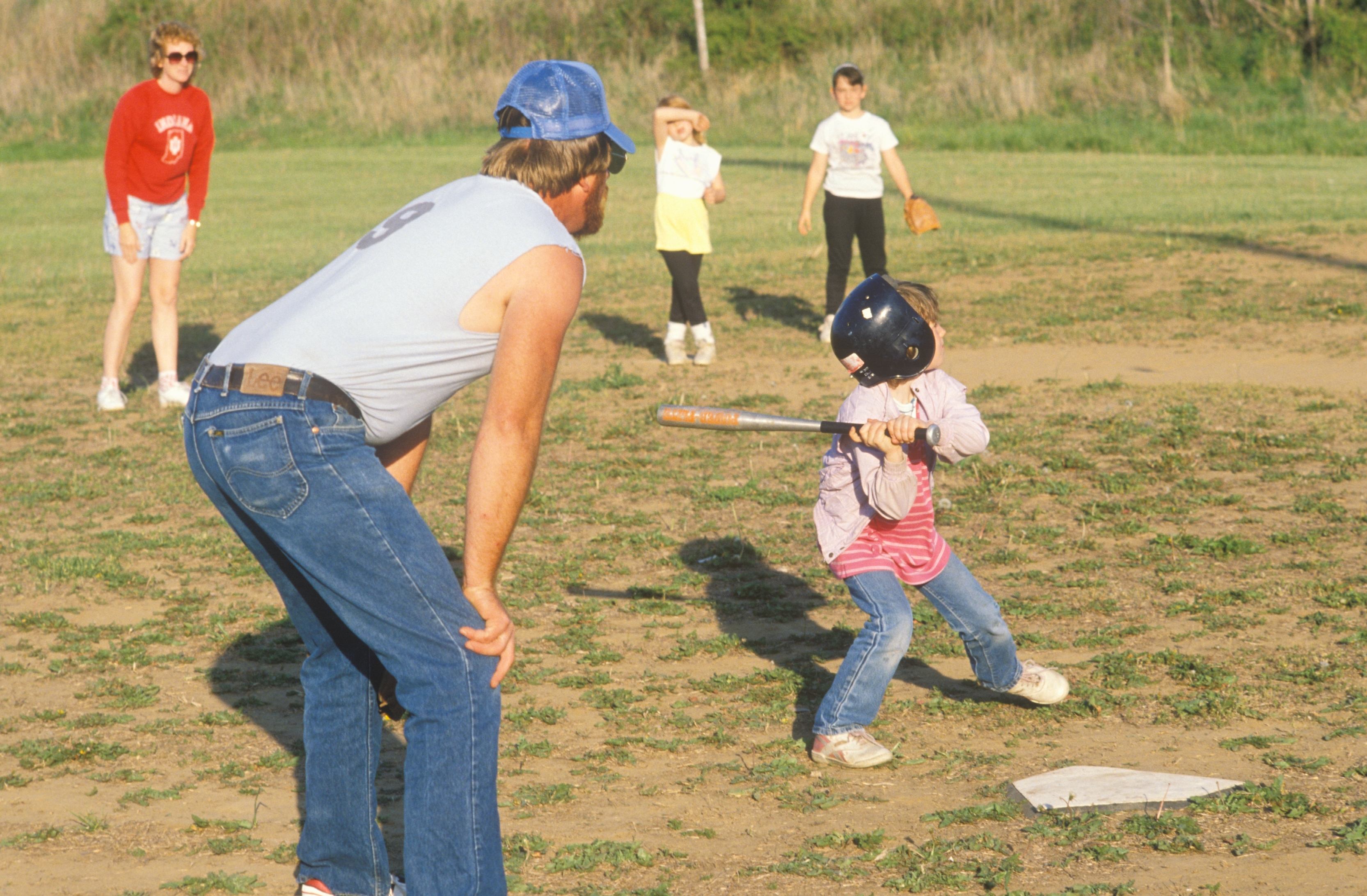 Child in a helmet and pink jacket prepares to swing bat during a casual baseball game, while an adult guides from nearby. Adults and kids watch in background