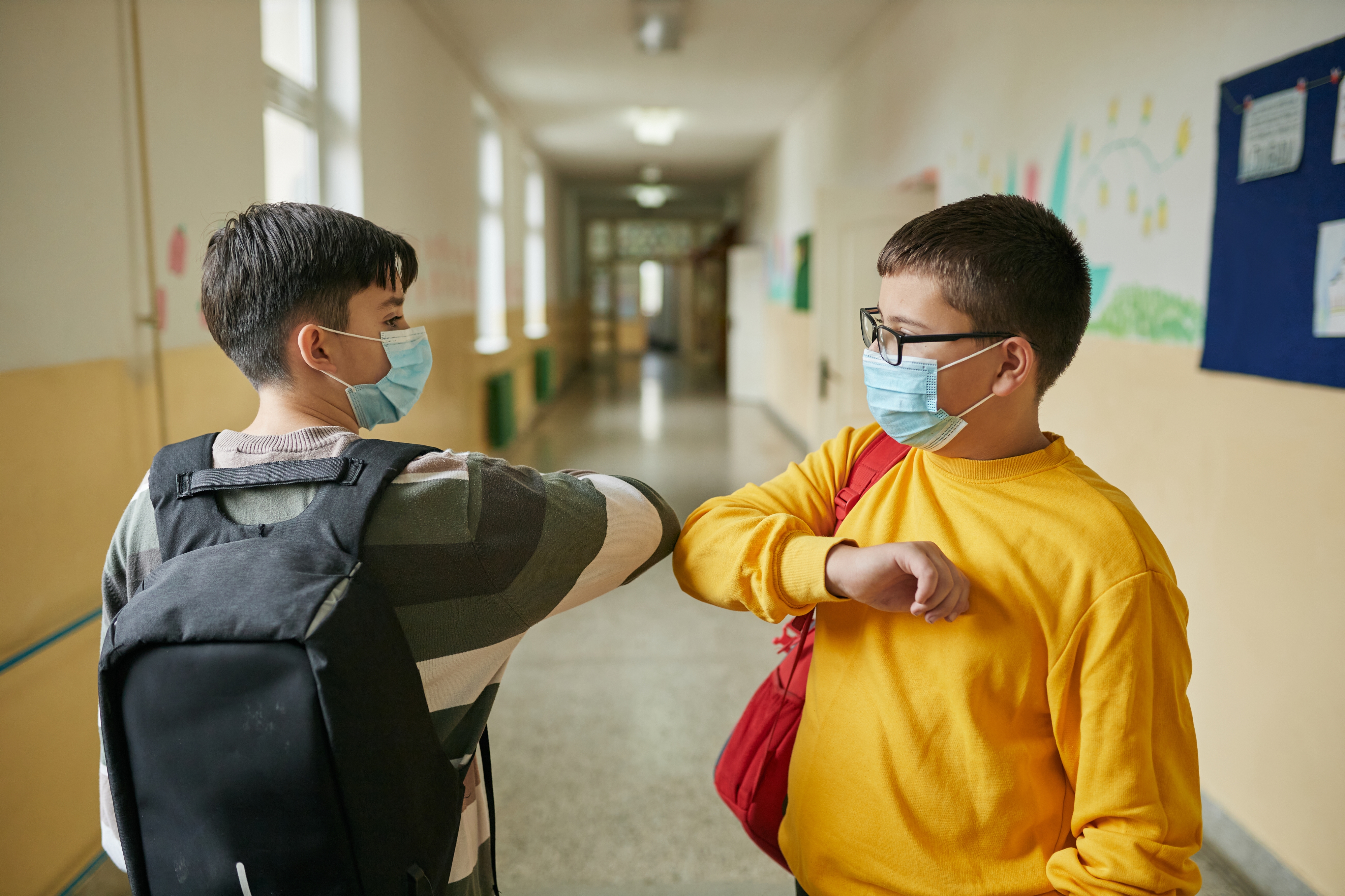 Two kids wearing masks and backpacks greet each other with elbow bumps in a school hallway