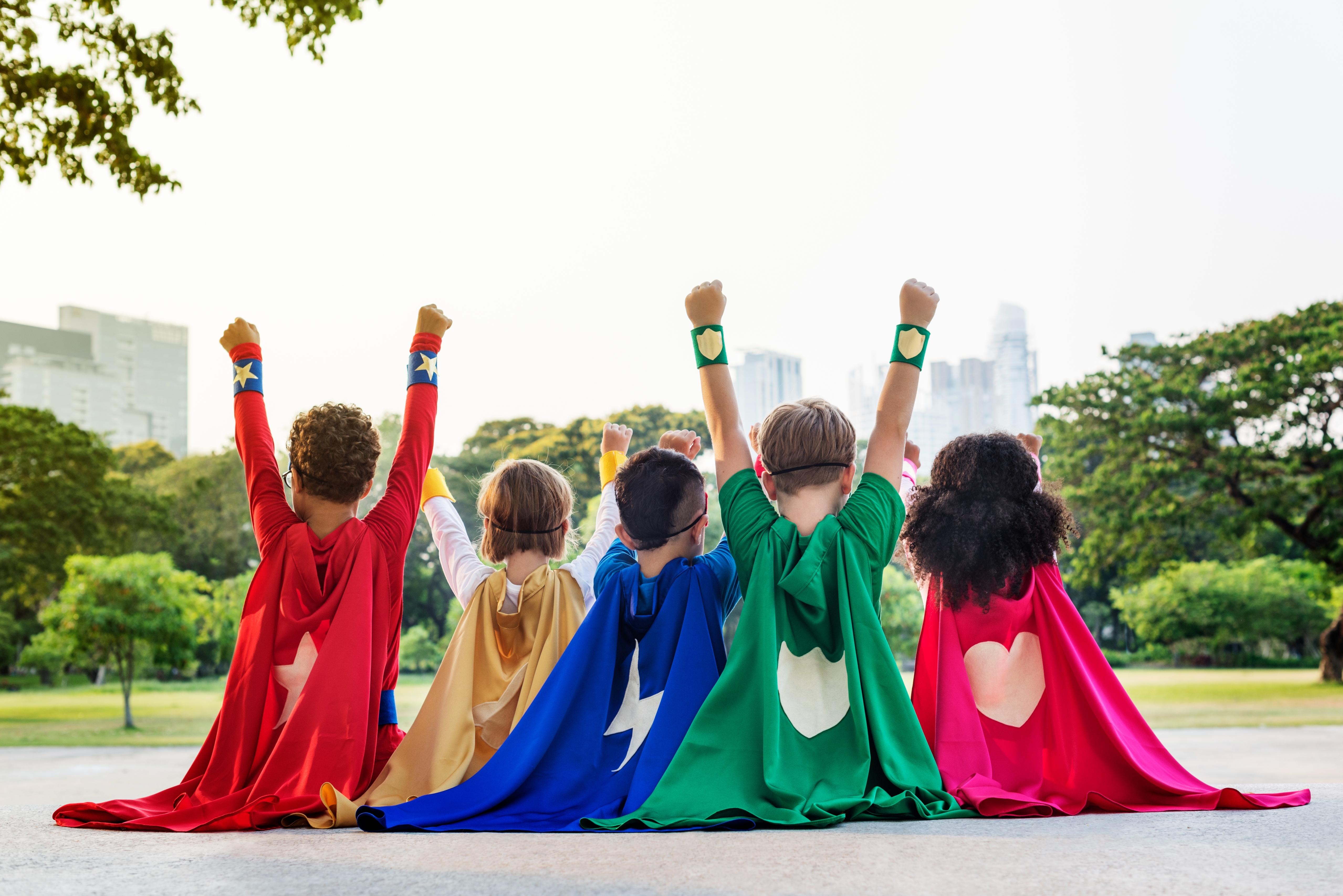 Children dressed as superheroes with capes sit on a grassy hill, raising their fists toward the city skyline in the background