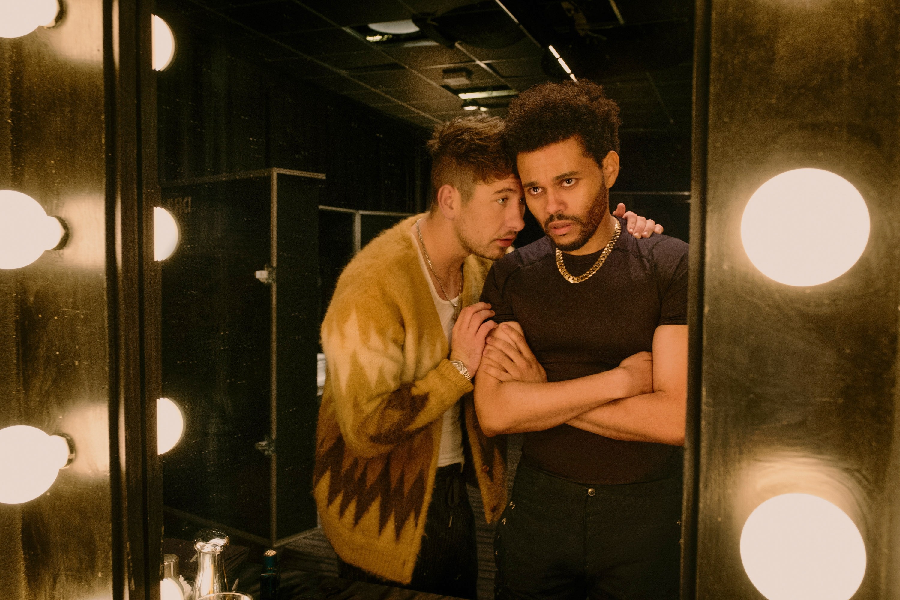 Two men in a dressing room. One wears a patterned cardigan and whispers to the other, who has a serious expression, wearing a black shirt and gold chain